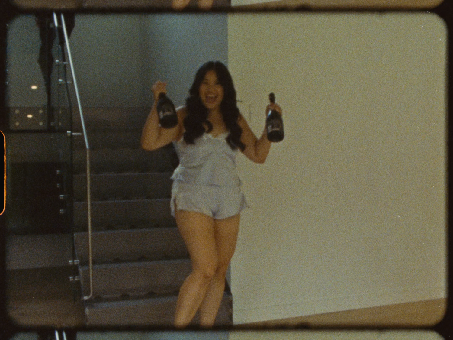 Woman in white shorts set holding two champagne bottles while descending a staircase.