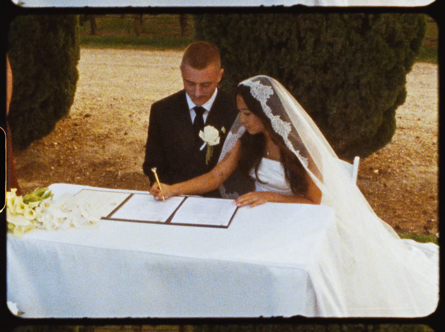 Bride and groom signing wedding registry documents at white draped table during outdoor ceremony.