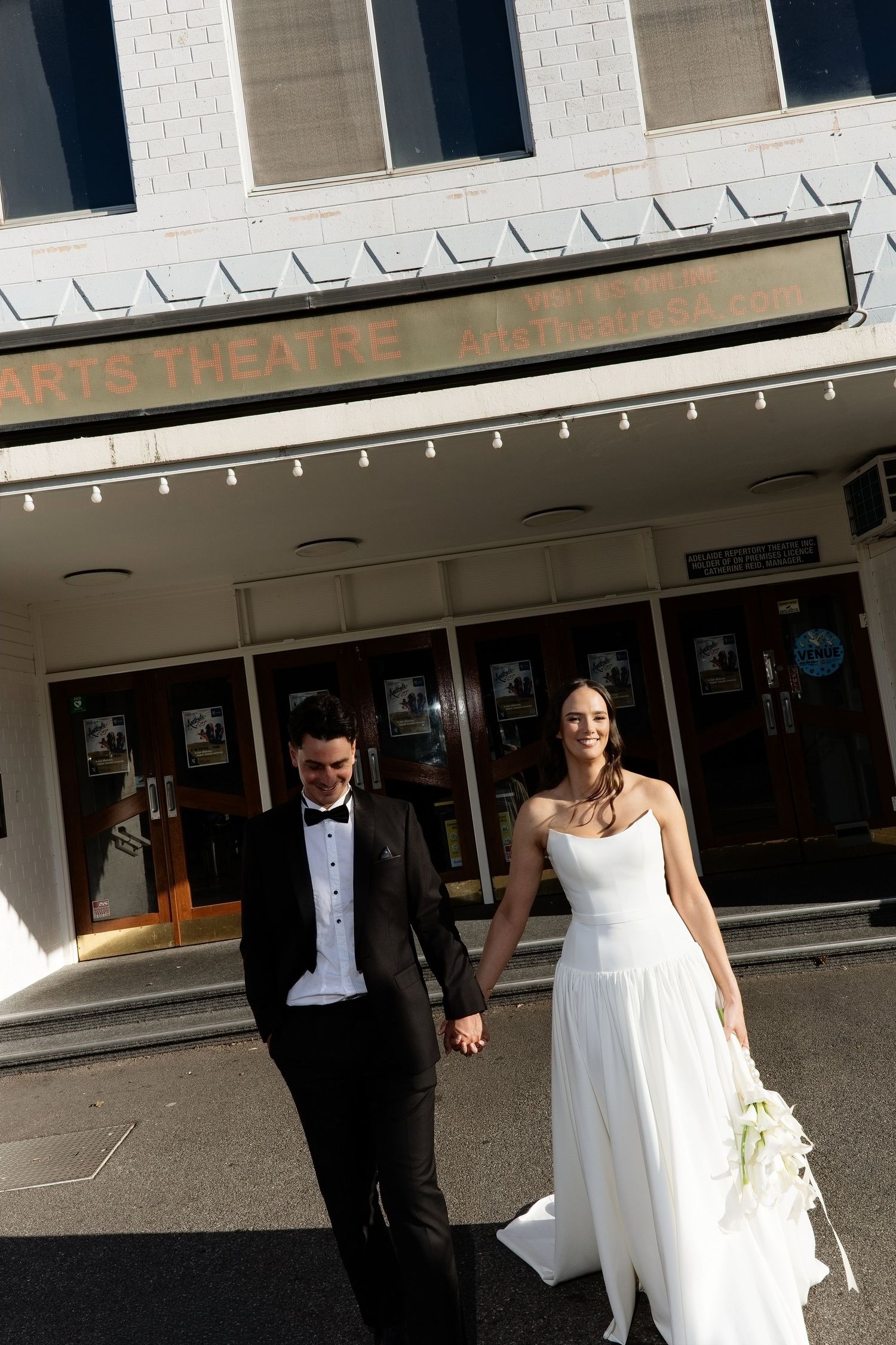 Elegant bride and groom seated before red doors, bride holding calla lily bouquet and pearl clutch bag.