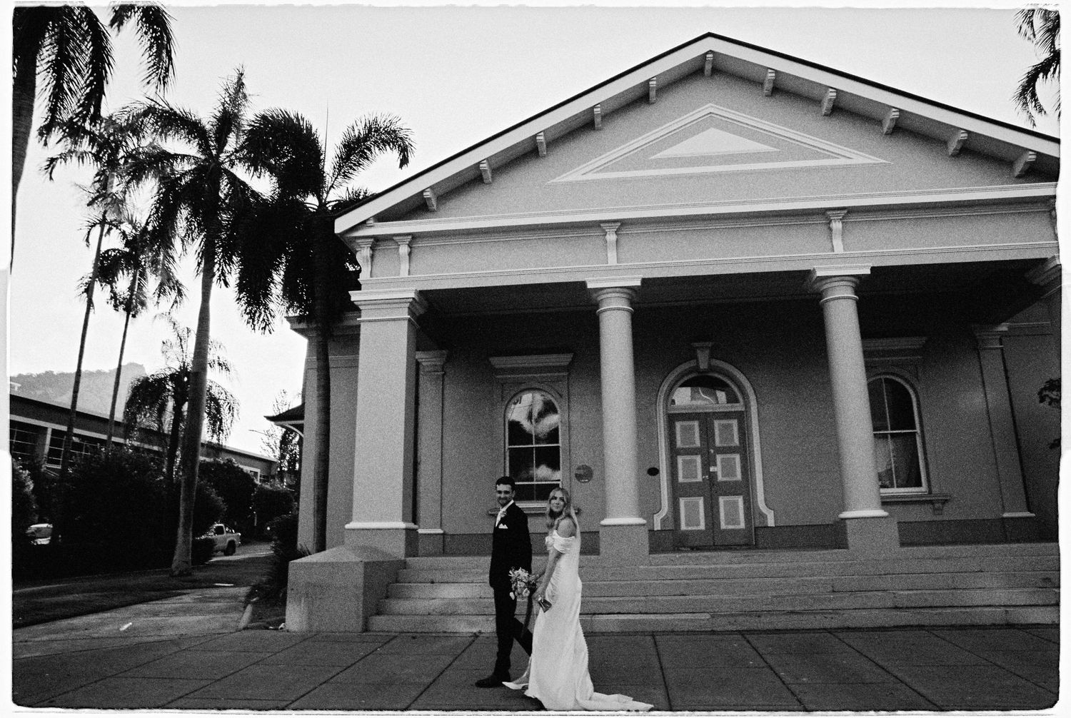 Black and white photo of a bride and groom posing outside a classic colonial-style building with palm trees.