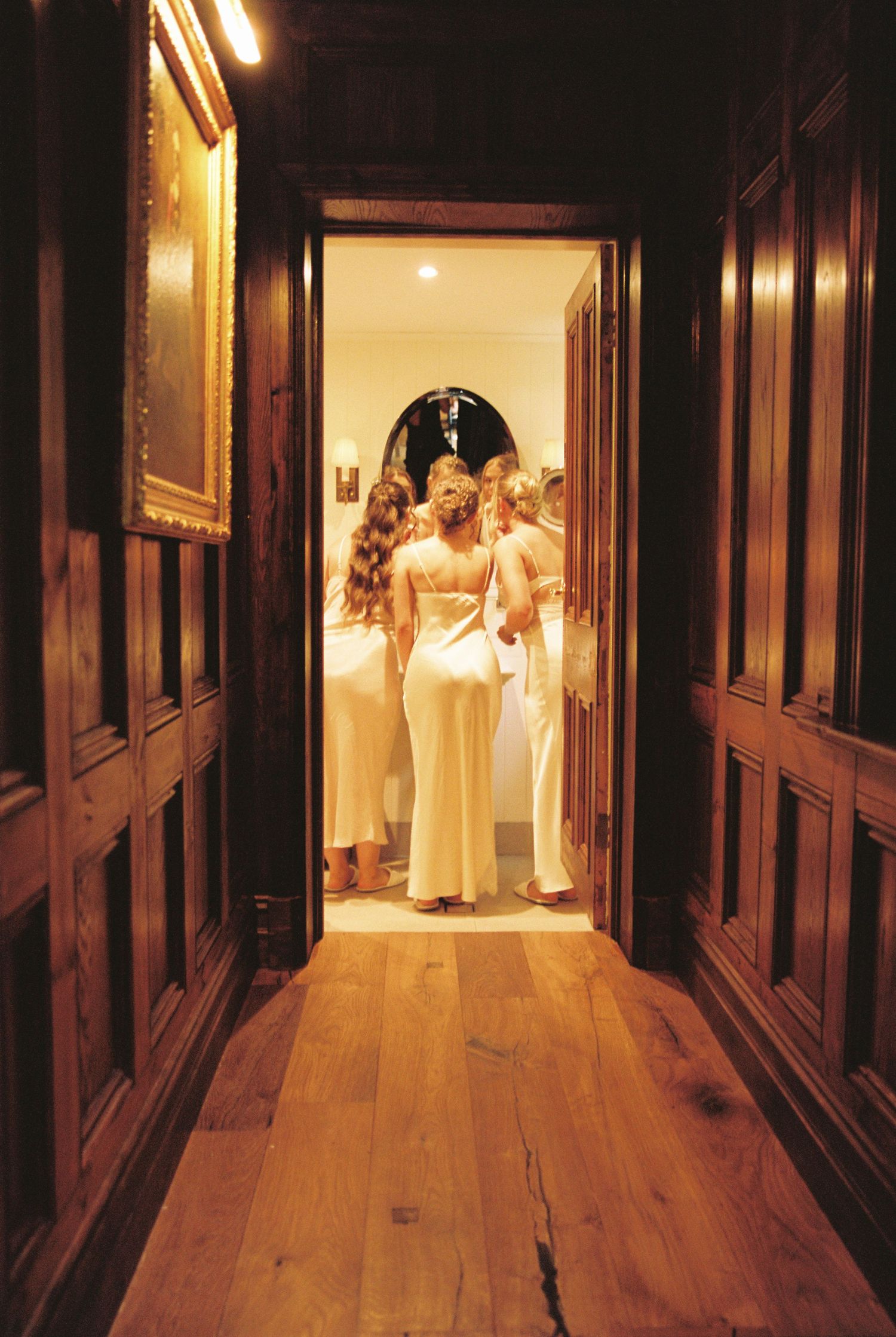 Bridesmaids in white gowns gathered in a warmly lit doorway at end of dark wood-paneled hallway.