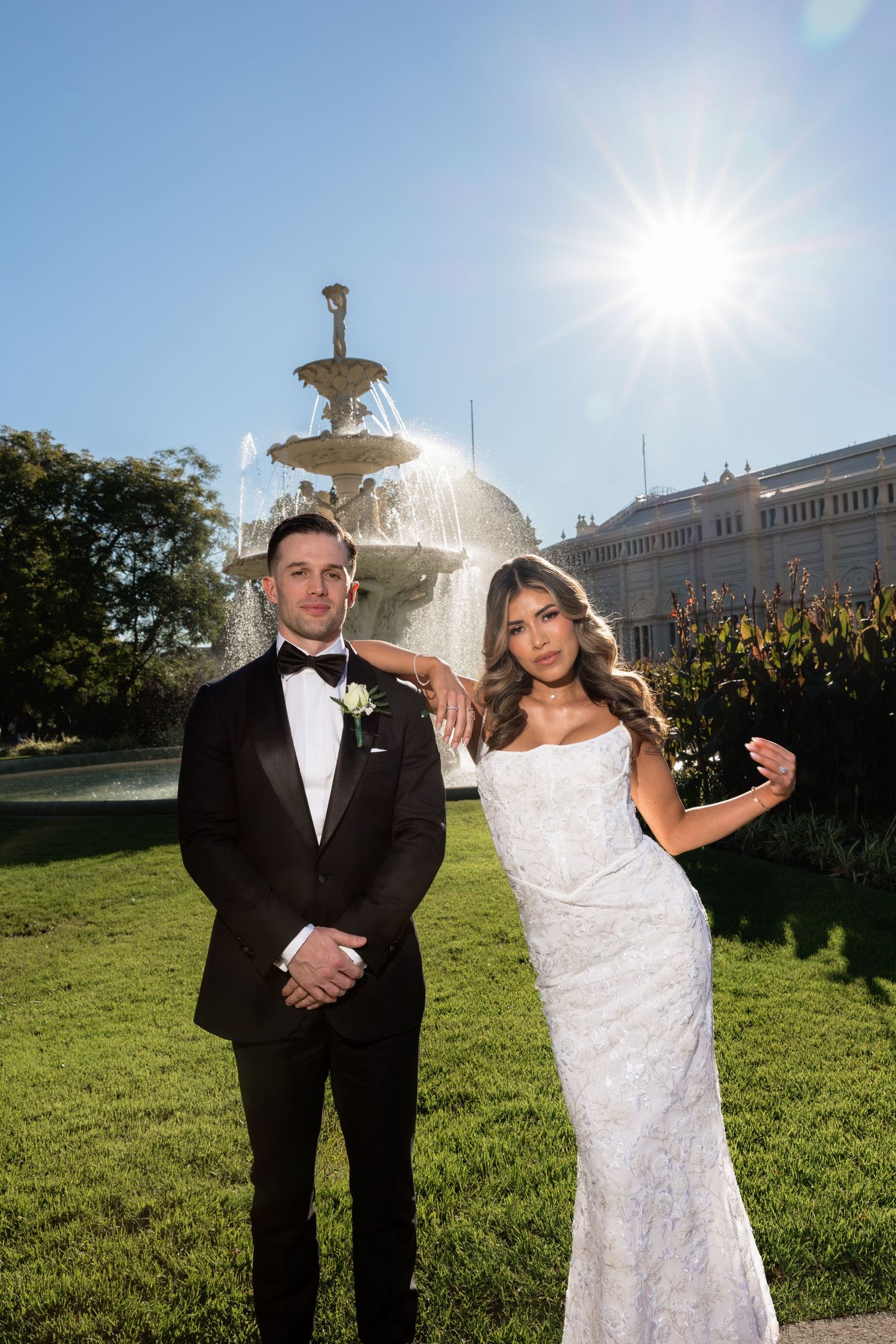 Bride and groom posing in front of a fountain on their wedding day in a sunny outdoor garden setting.