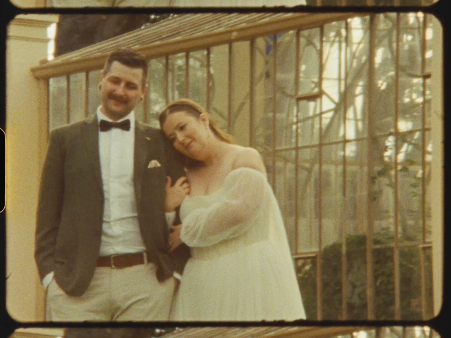 Smiling bride and groom posing together in vintage film style before ornate iron garden gates.