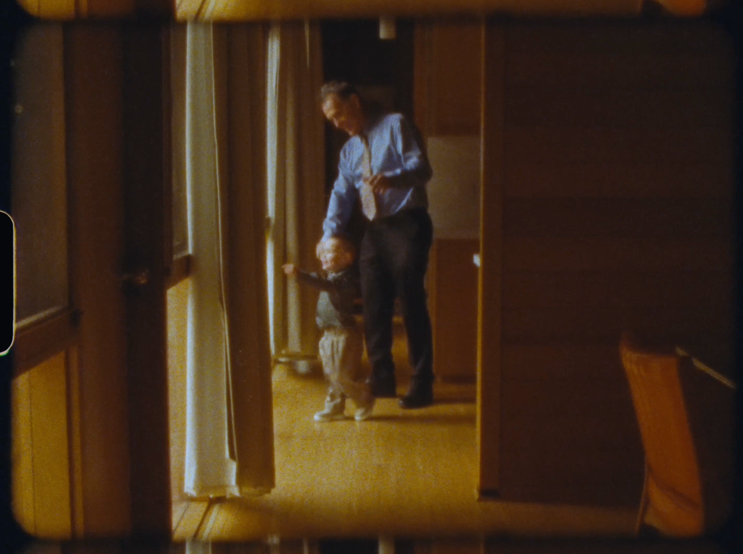 An adult helps a toddler walk in a warmly lit room with wooden walls.