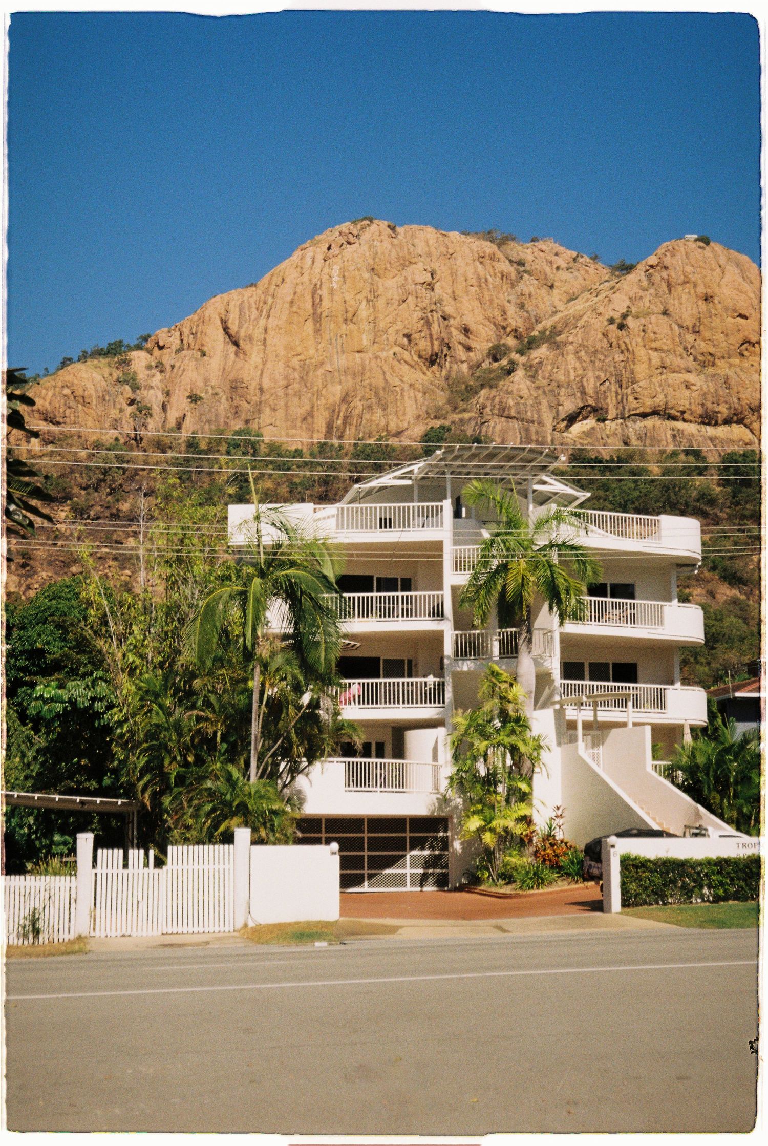White multi-story apartment building with palm trees backdropped by a large rocky mountain under a clear blue sky.