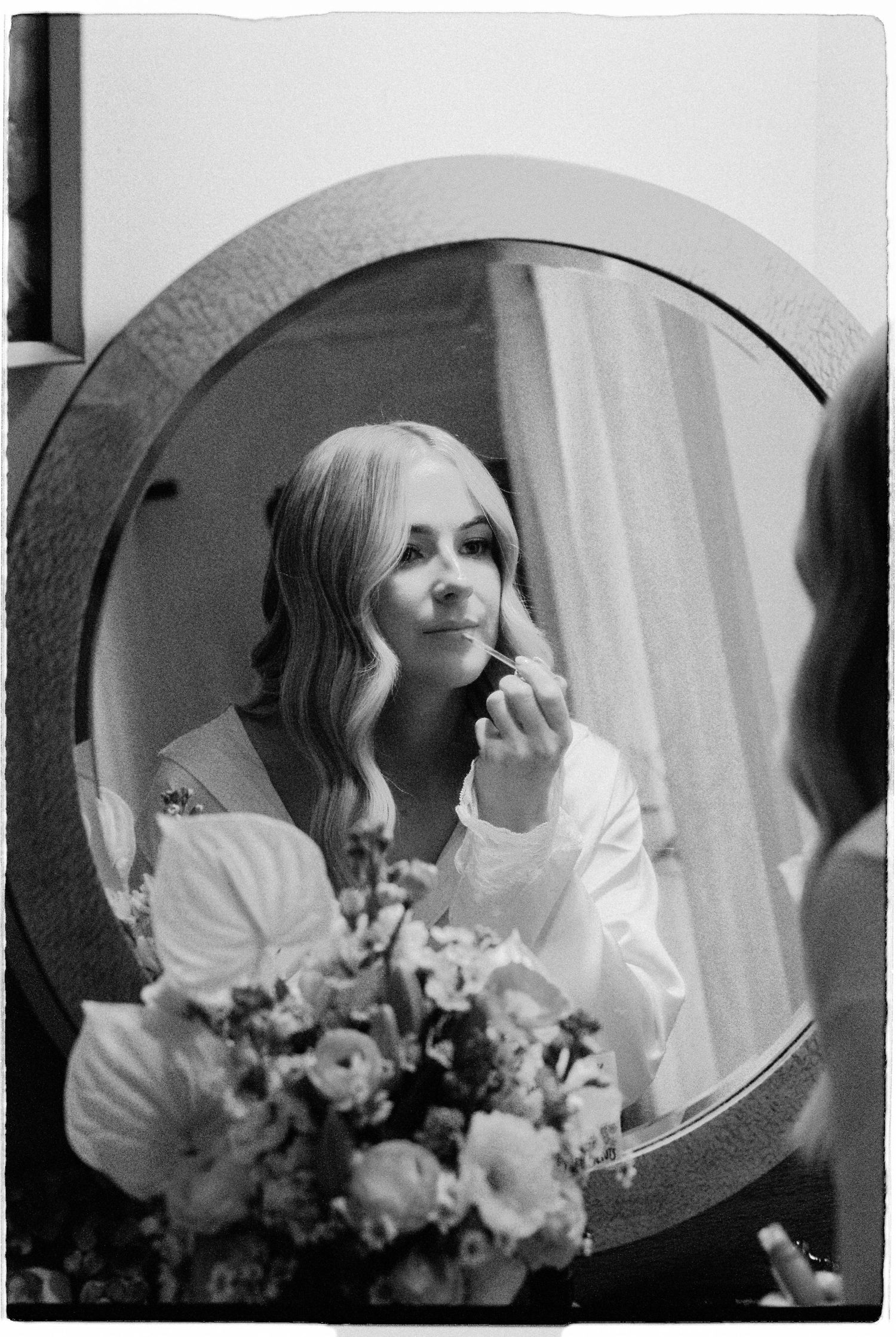 Black and white photo of bride applying makeup in round mirror with bridal bouquet below.
