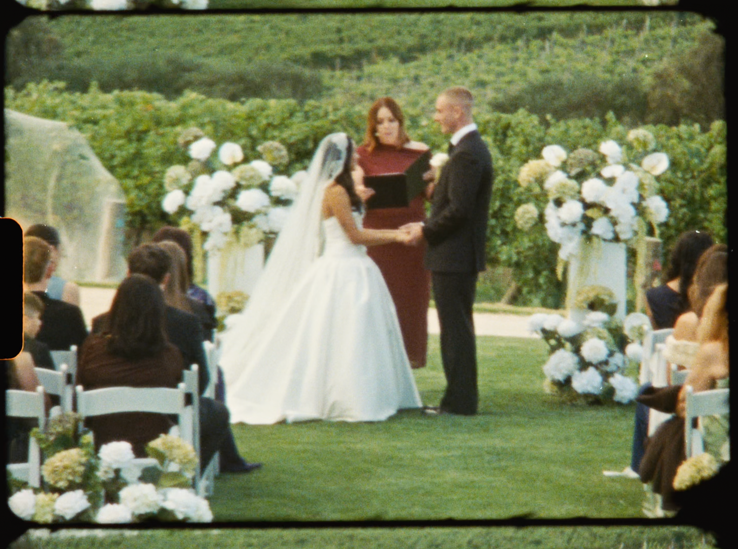 Bride and groom exchanging vows at elegant outdoor vineyard wedding with white floral arch.