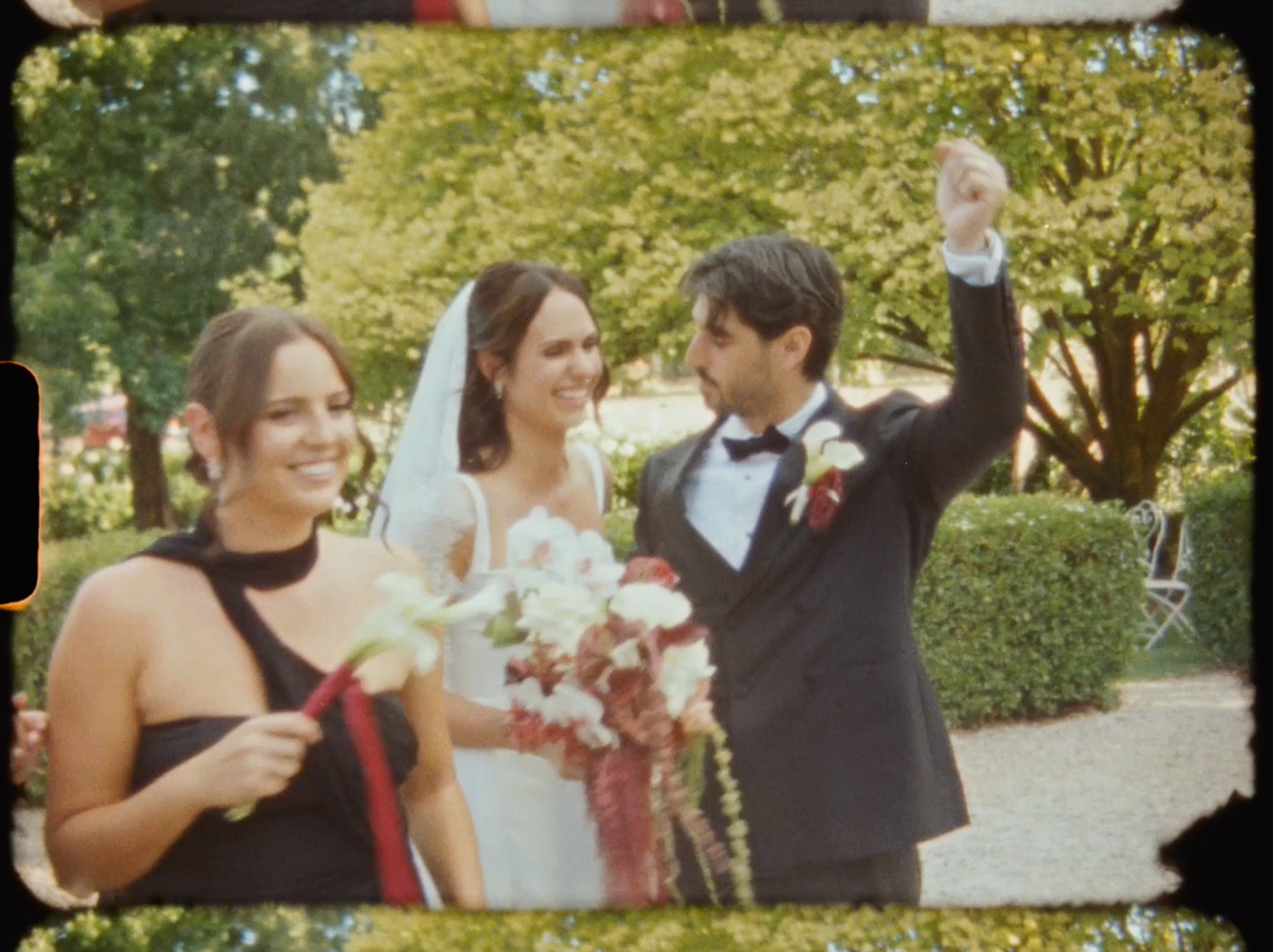 Joyful bride and groom celebrating with a raised fist alongside a bridesmaid in a lush garden setting.