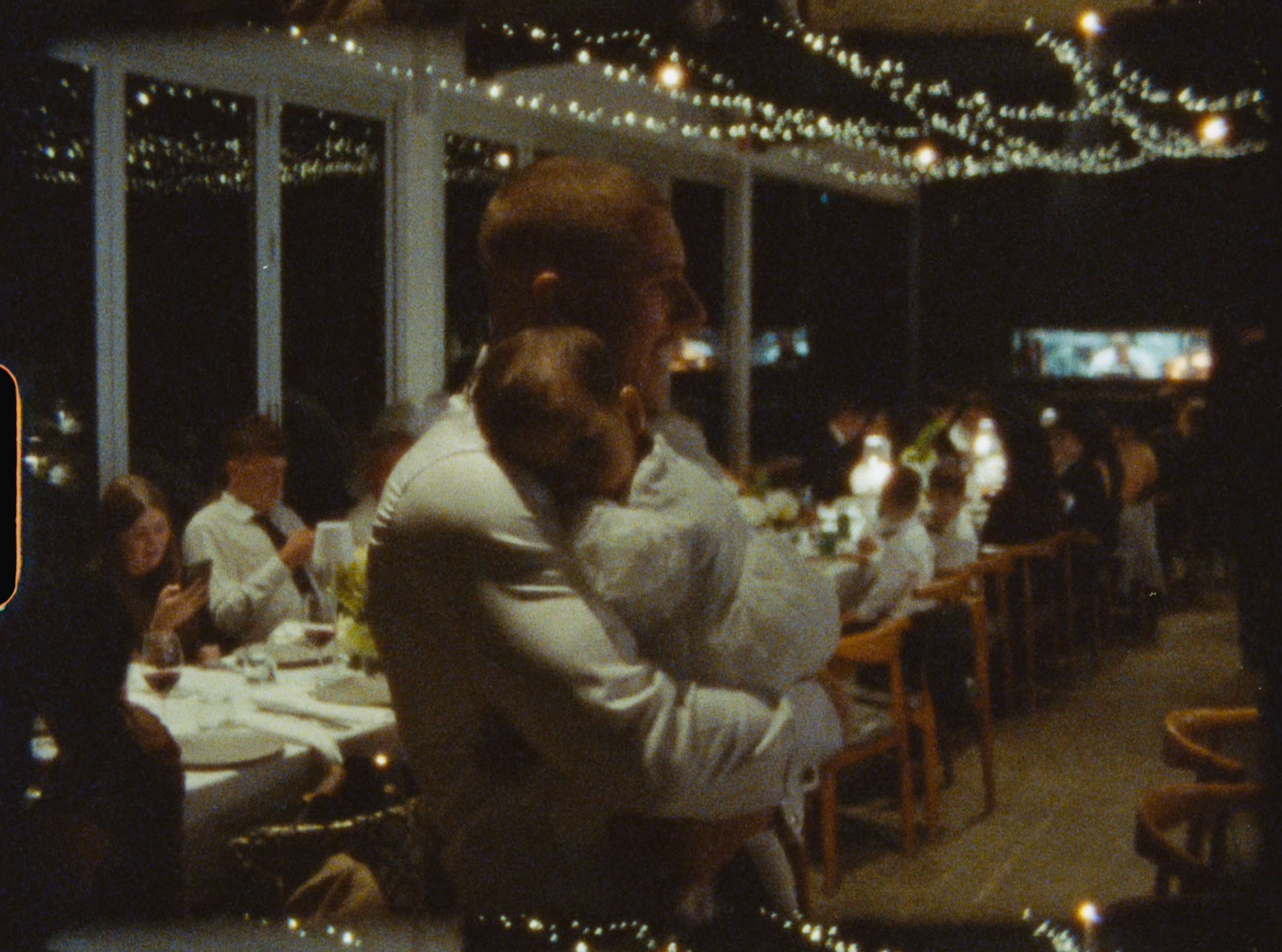 Wedding guests seated at outdoor reception tables under warm string lights at night.