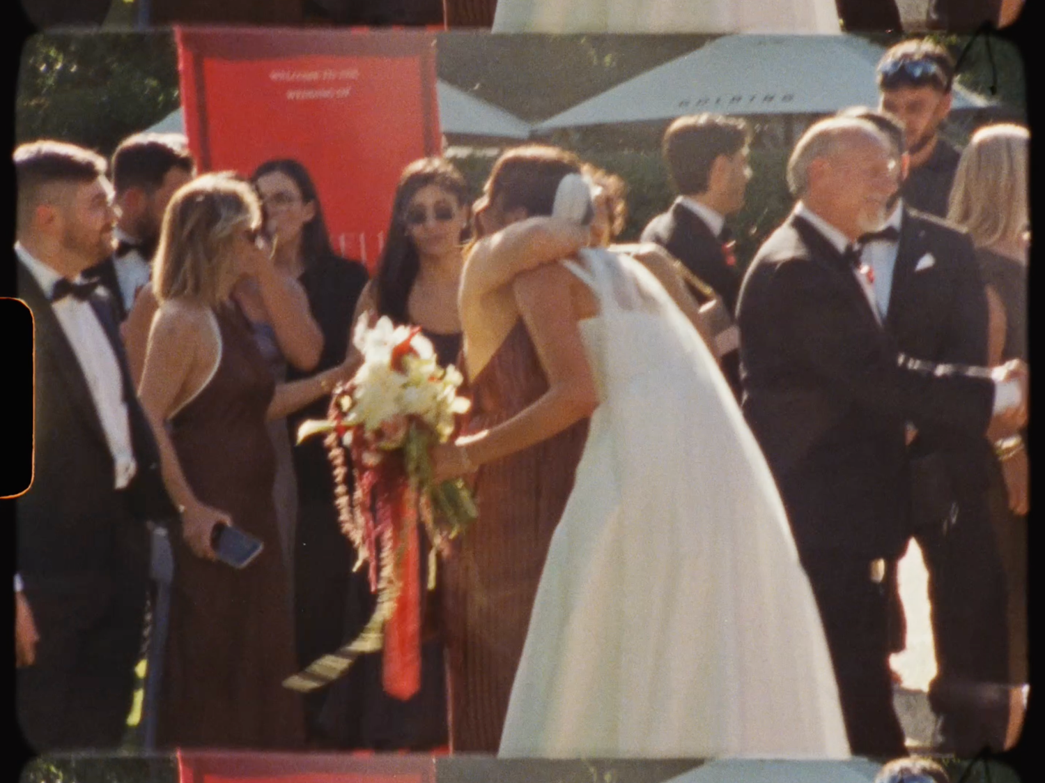 Bride in a white gown embraces a guest while holding a colorful bridal bouquet at outdoor reception.
