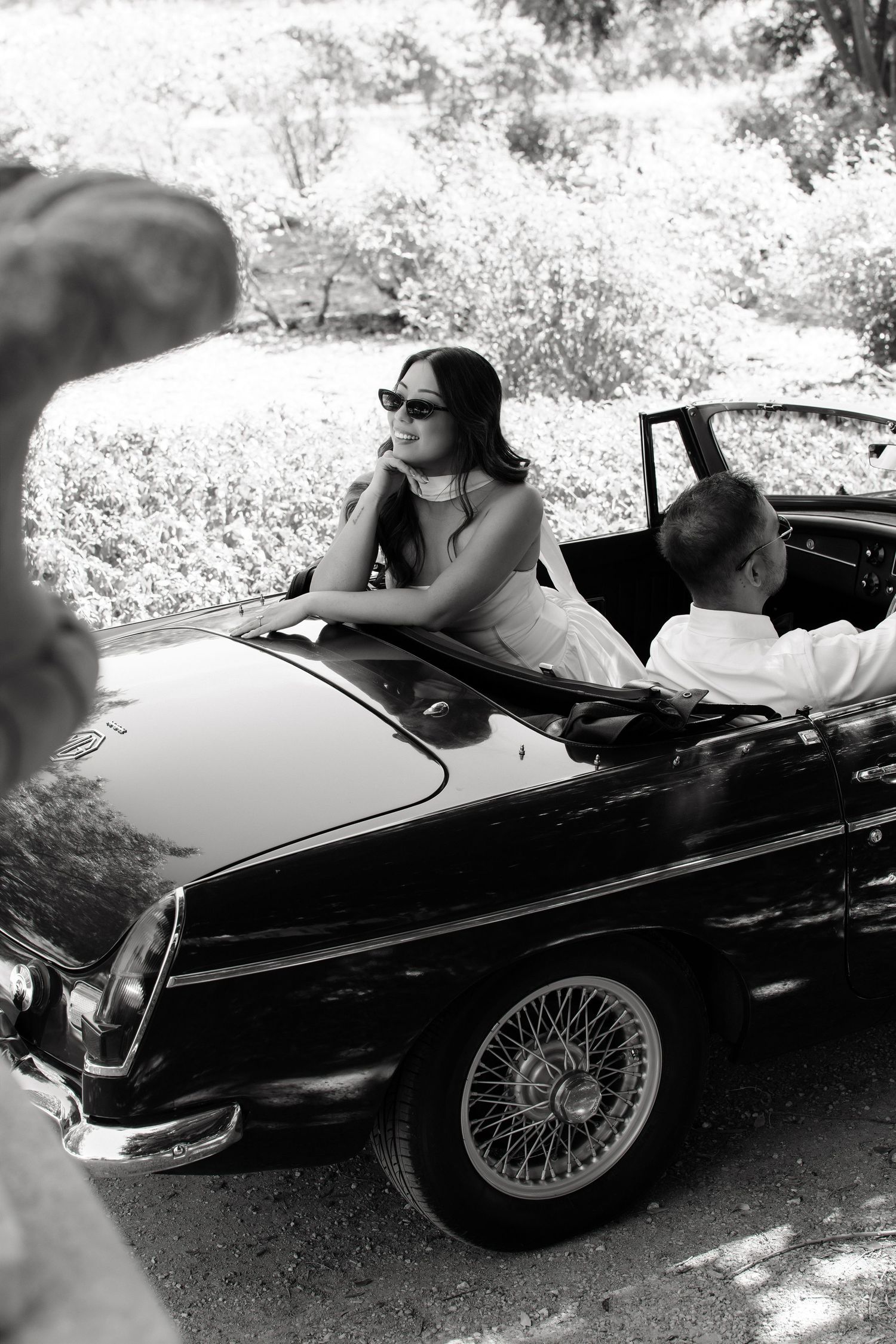 Bride in sunglasses posing in vintage convertible car in black and white photo.