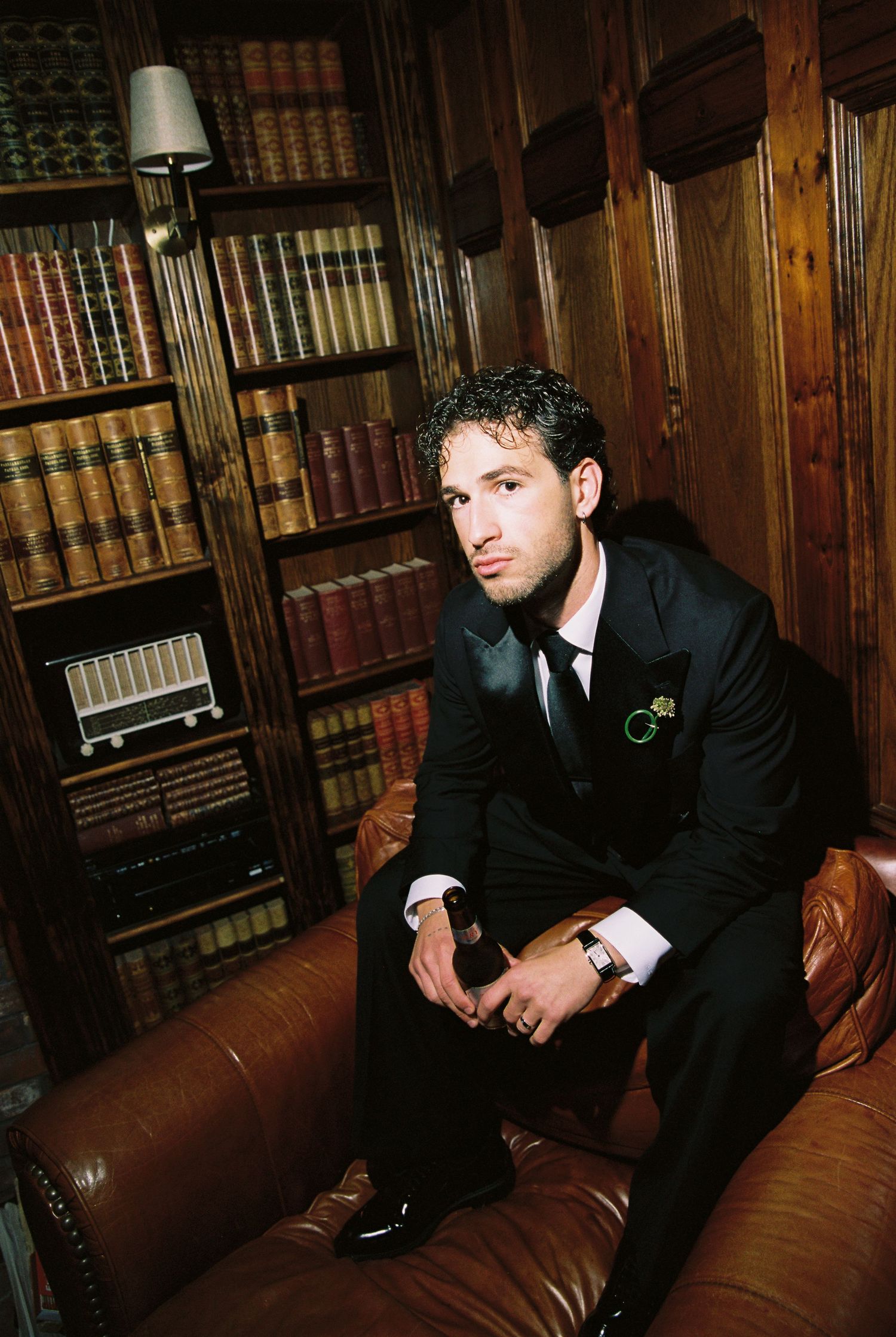 Groom in black suit seated on leather sofa in wood-paneled library with vintage books in background.