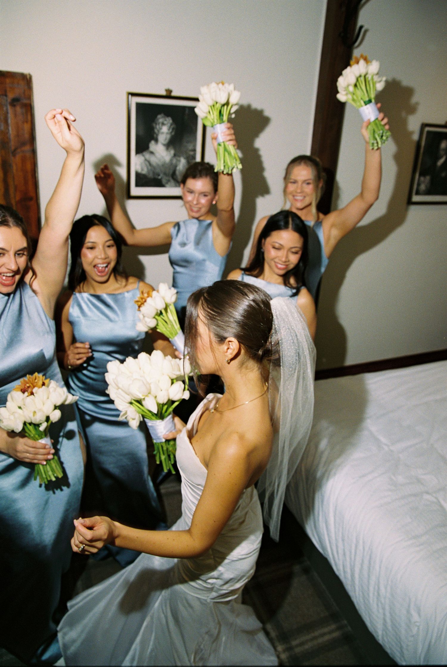 Excited bridesmaids in blue satin dresses cheering with white tulip bouquets around a bride on her wedding day.