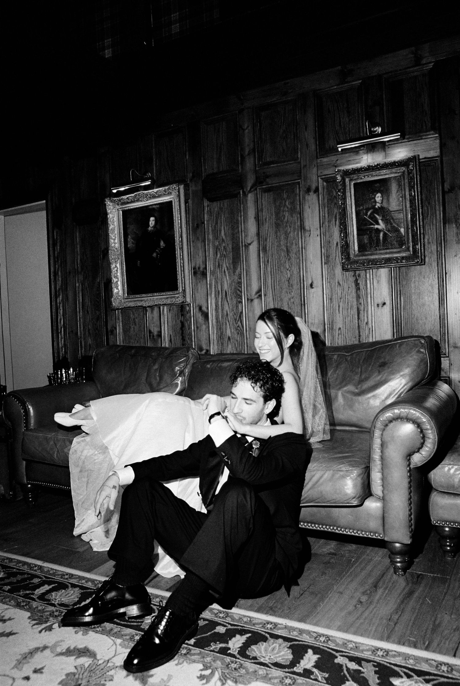 Black and white photo of bride and groom sitting together on leather sofa with portrait paintings on wood-paneled wall.