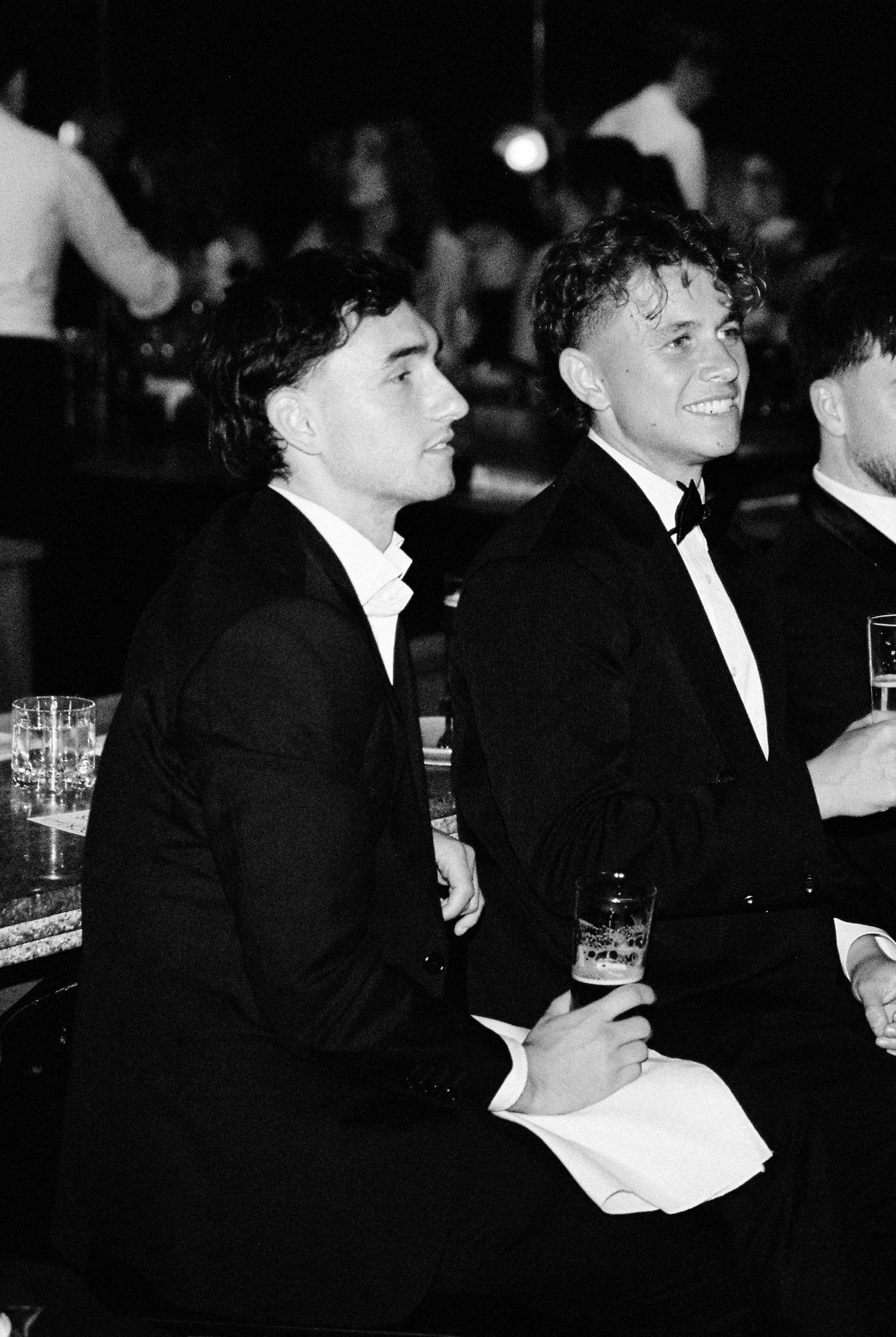 Two young men in tuxedos sit together smiling at wedding reception in black and white photo.