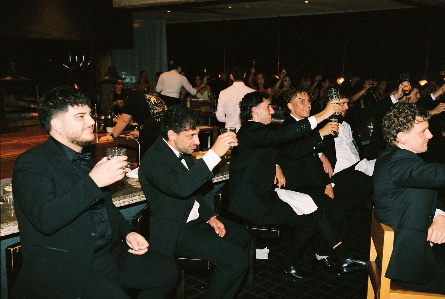 Groomsmen in black tuxedos raising glasses in a toast at wedding reception.