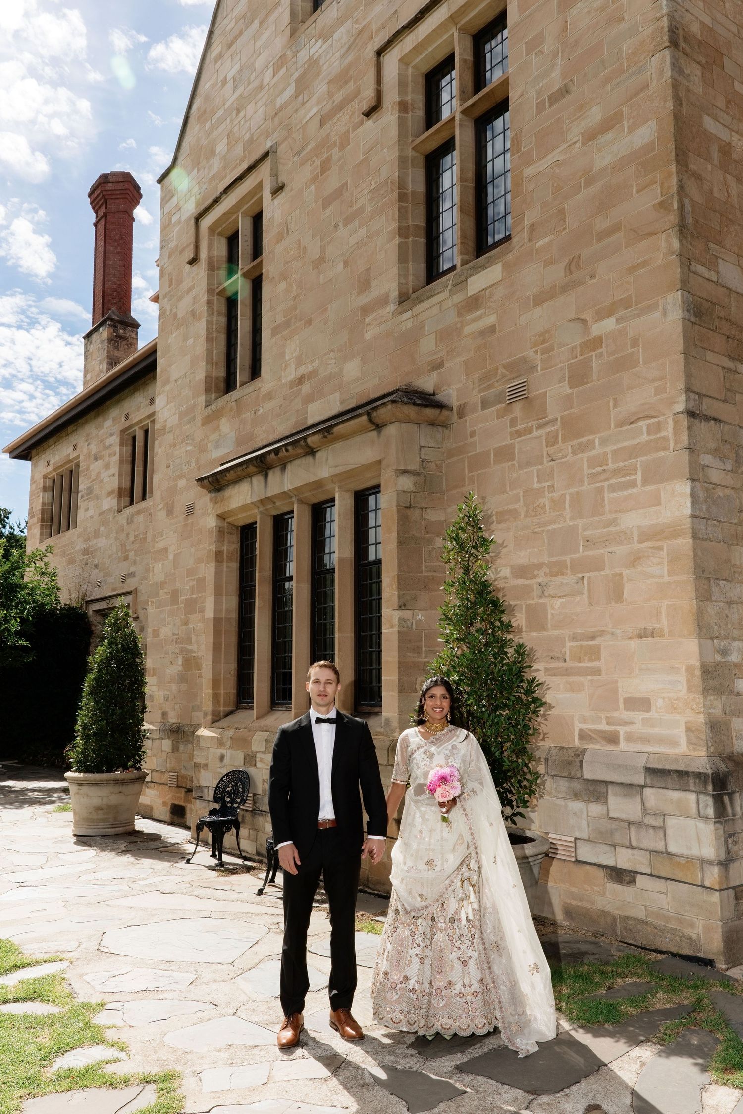 Bride in Indian bridal lehenga and groom in tuxedo standing before a historic sandstone building.