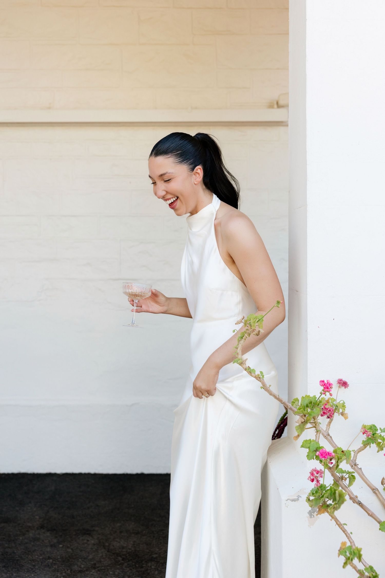 Elegant bride in white satin halter dress laughs while holding champagne glass beside pink flowers outdoors.
