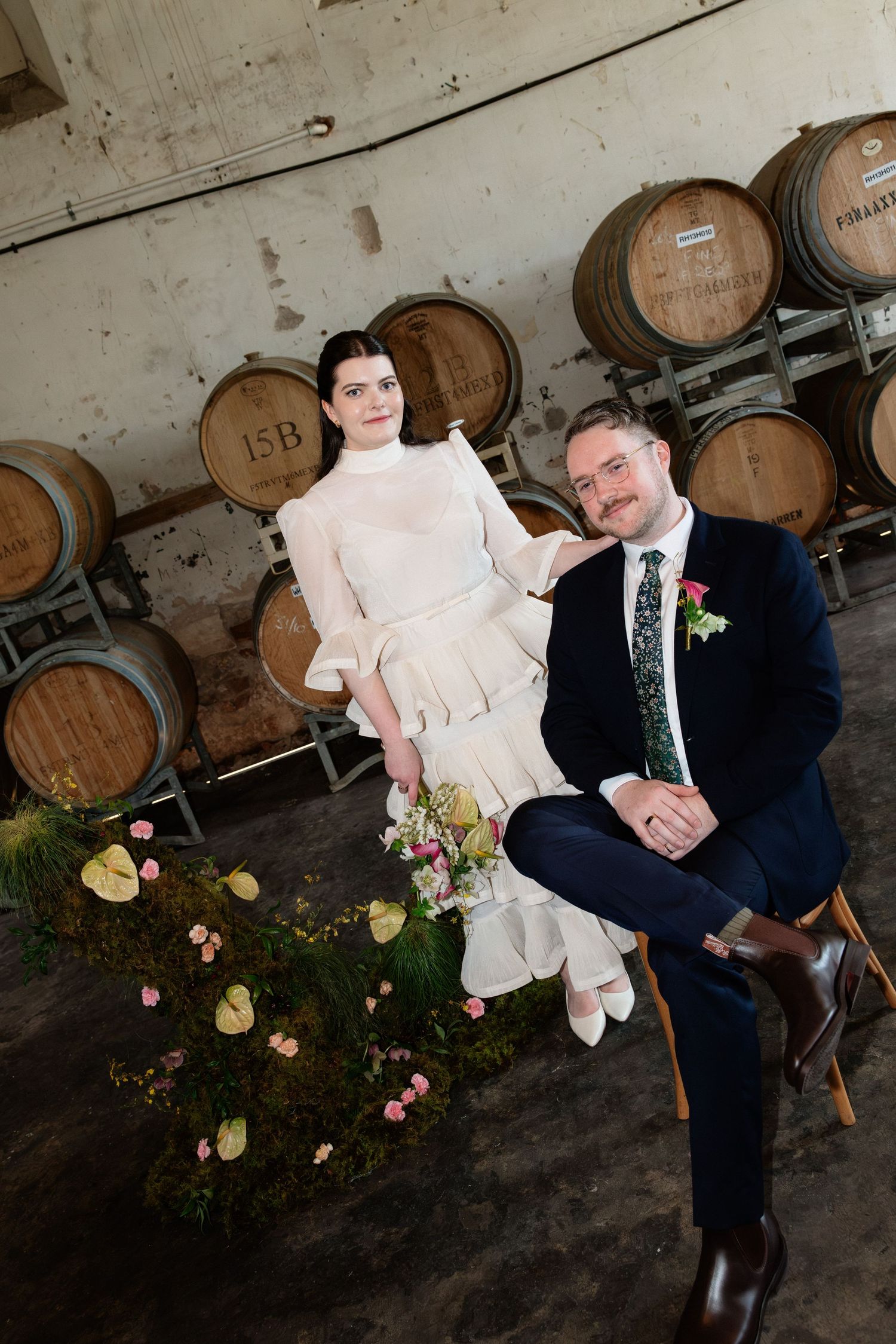 Bride and groom pose together among oak wine barrels with a romantic floral moss arrangement at their winery wedding.
