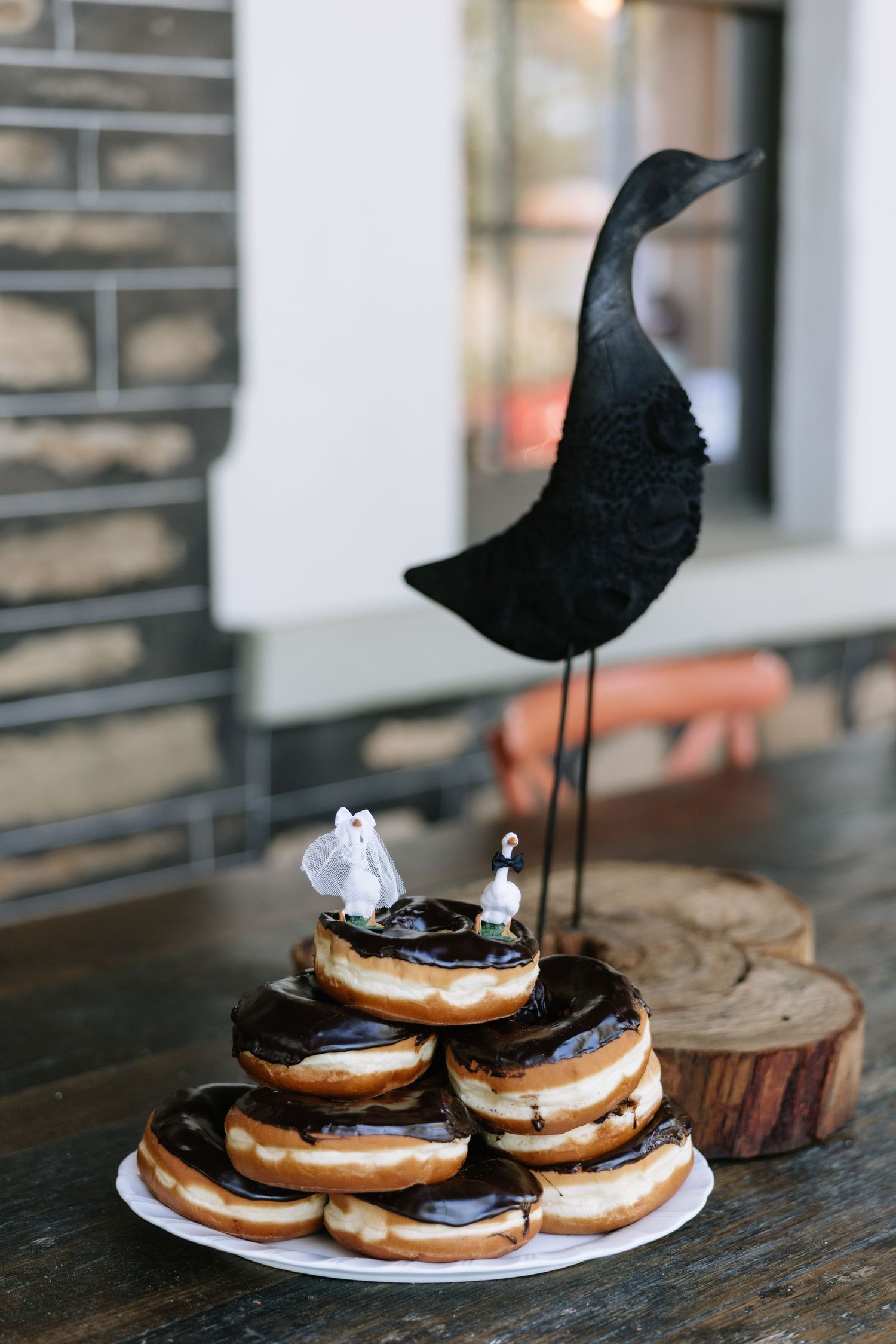 Stacked chocolate glazed donuts topped with bride and groom figurines and black bird sculpture on wooden table.