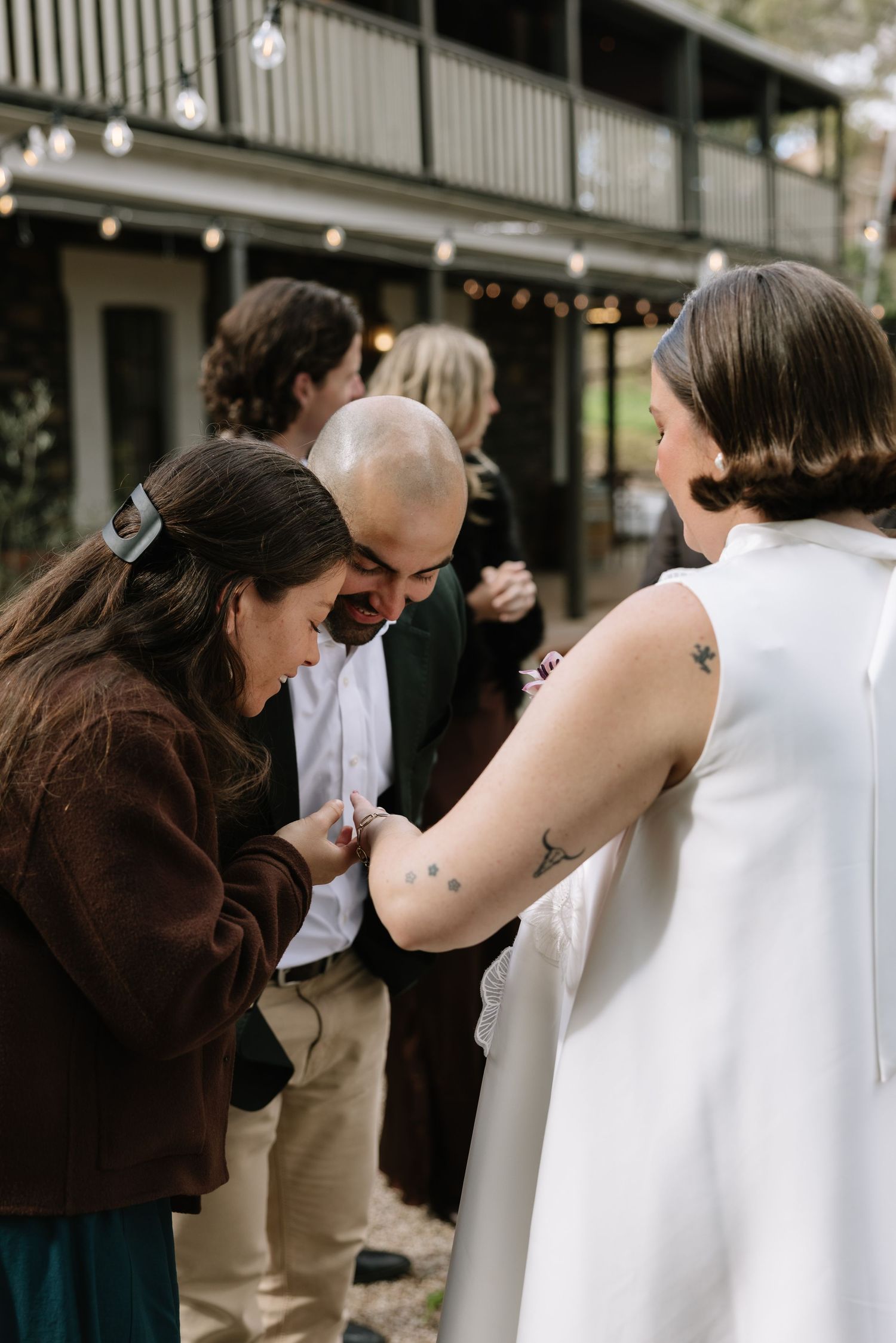 Guests admire a tattoo on a bride's arm at an outdoor wedding reception with string lights.