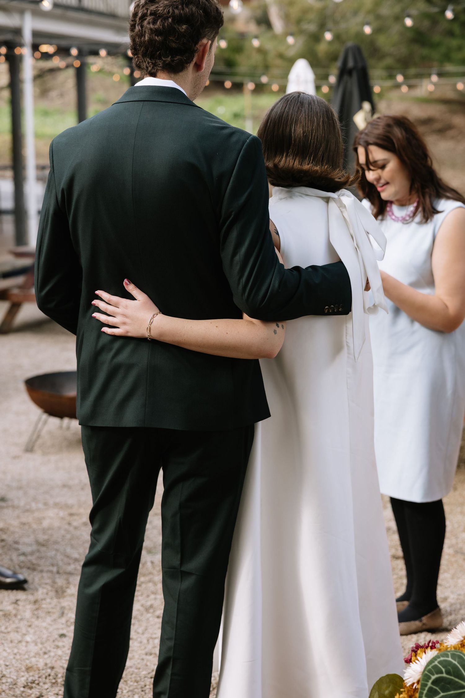 Bride and groom embrace during outdoor wedding ceremony while officiant looks on, string lights in background.