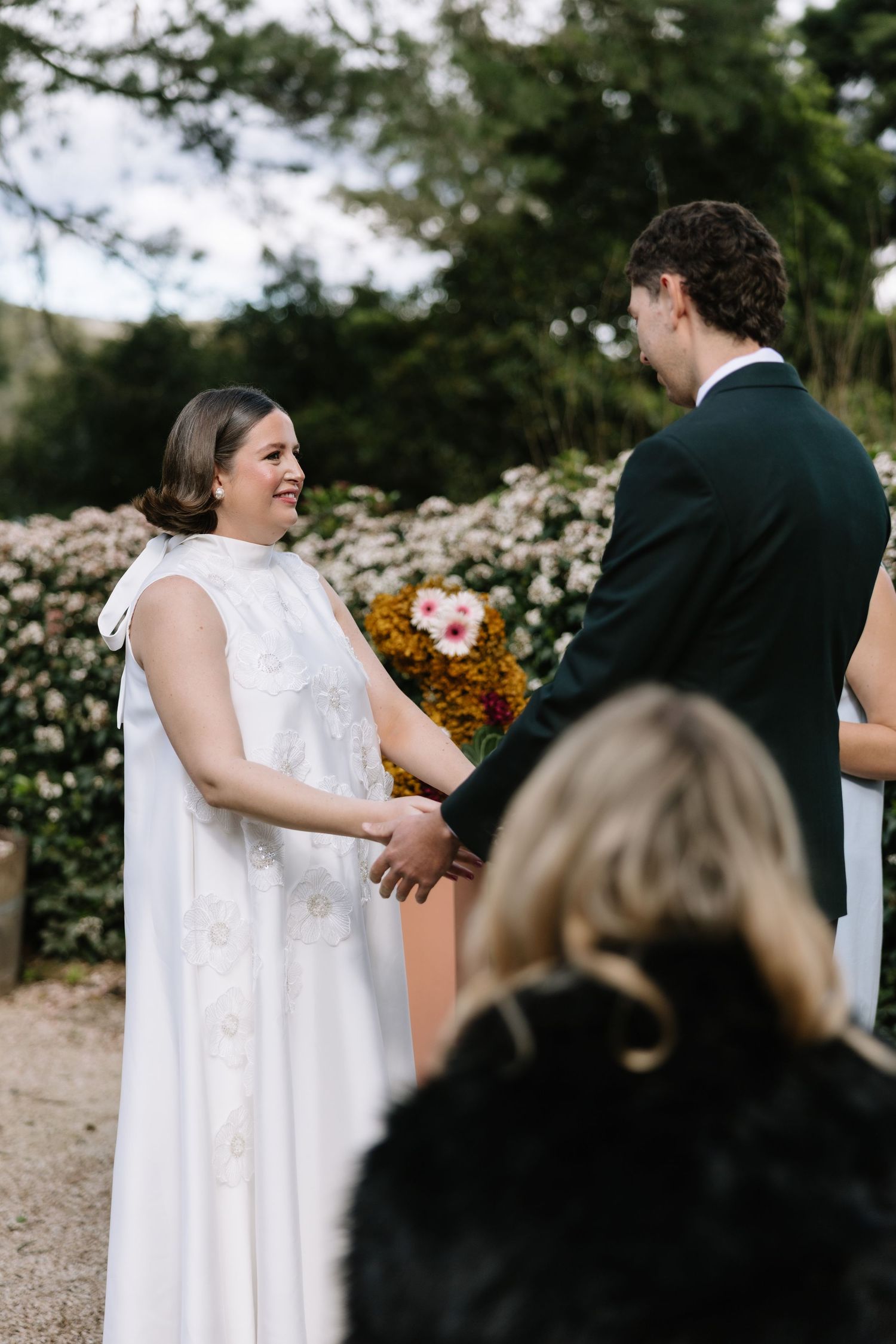 Bride in white gown holds hands with groom in dark suit during outdoor wedding ceremony surrounded by blooming flowers.