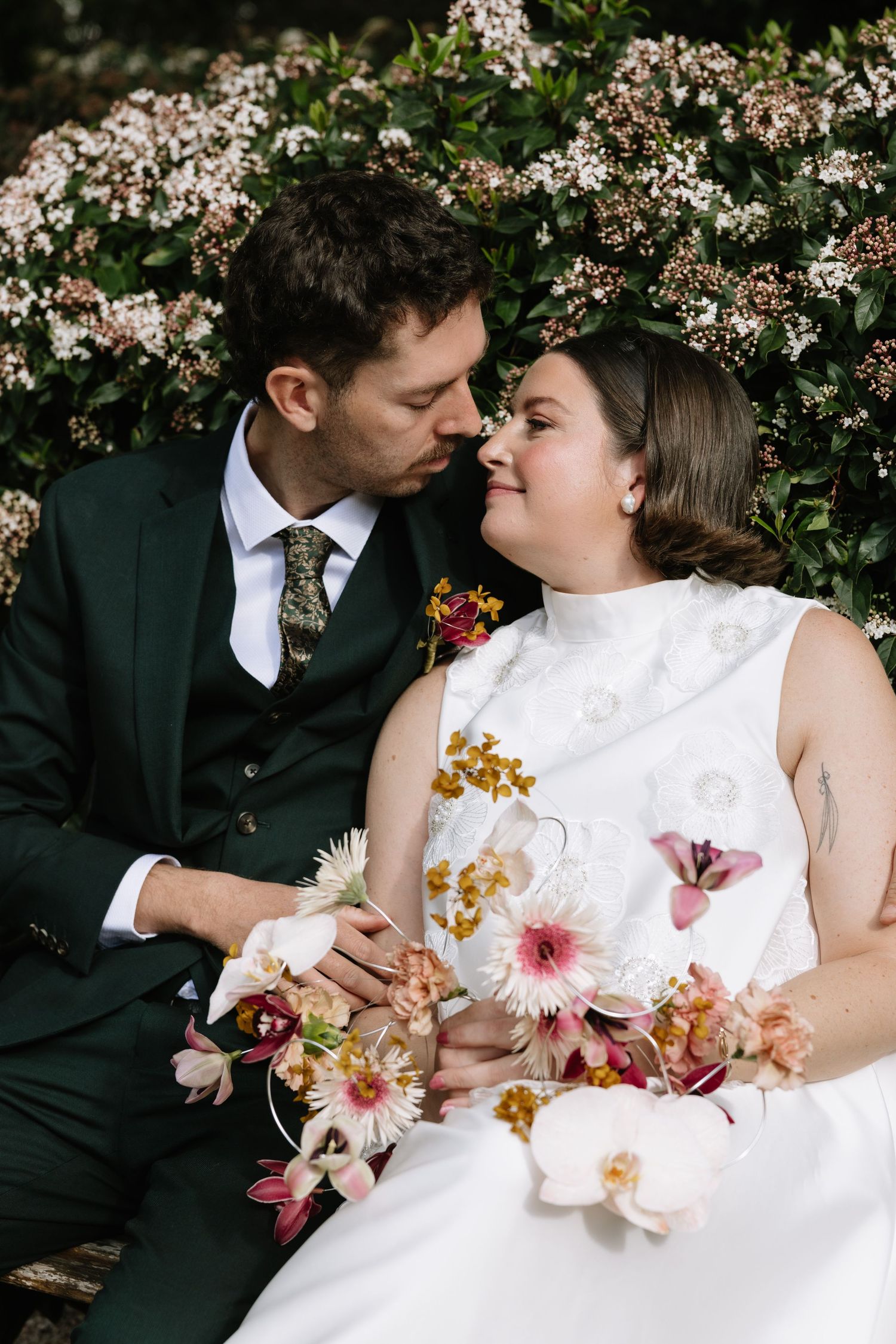 A bride and groom share a romantic moment surrounded by blooming flowers, she holds a colorful bouquet.