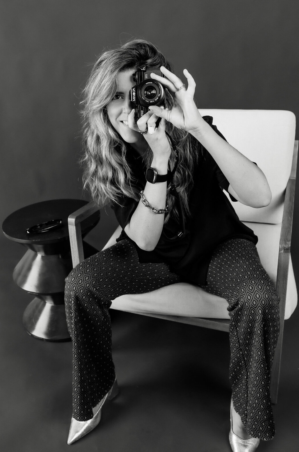 Black and white photo of a woman with wavy hair sitting in a white chair, holding a camera up to her face.