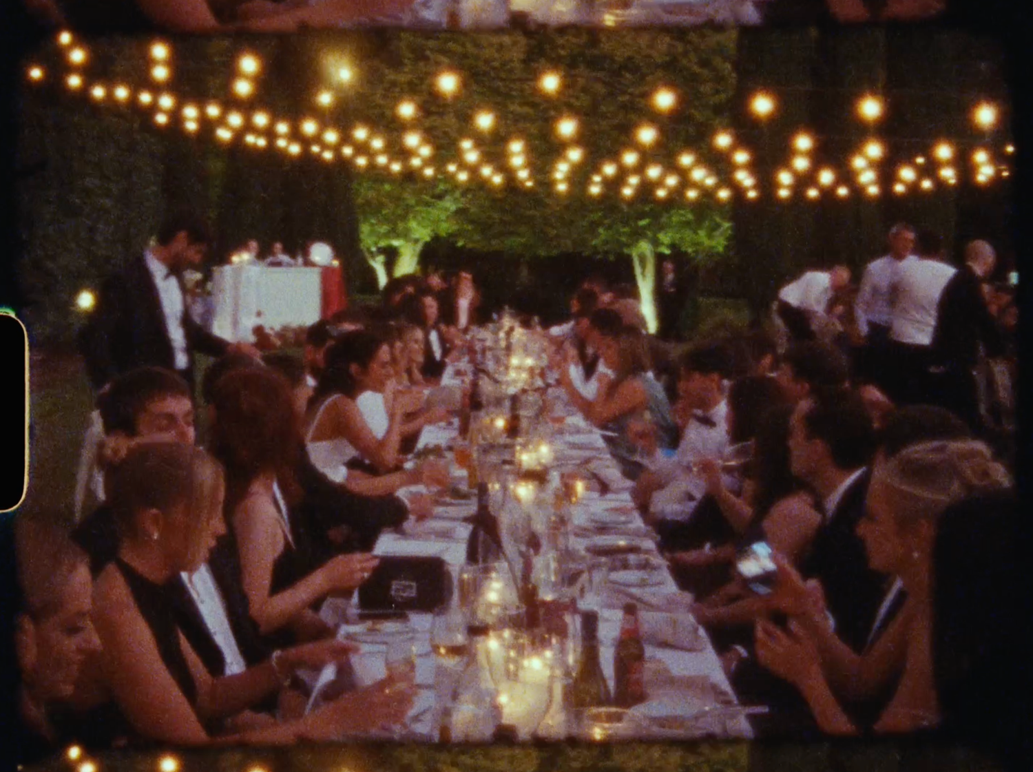 Elegant outdoor wedding reception dinner with string lights, candles, and guests in formal attire at a long decorated table.