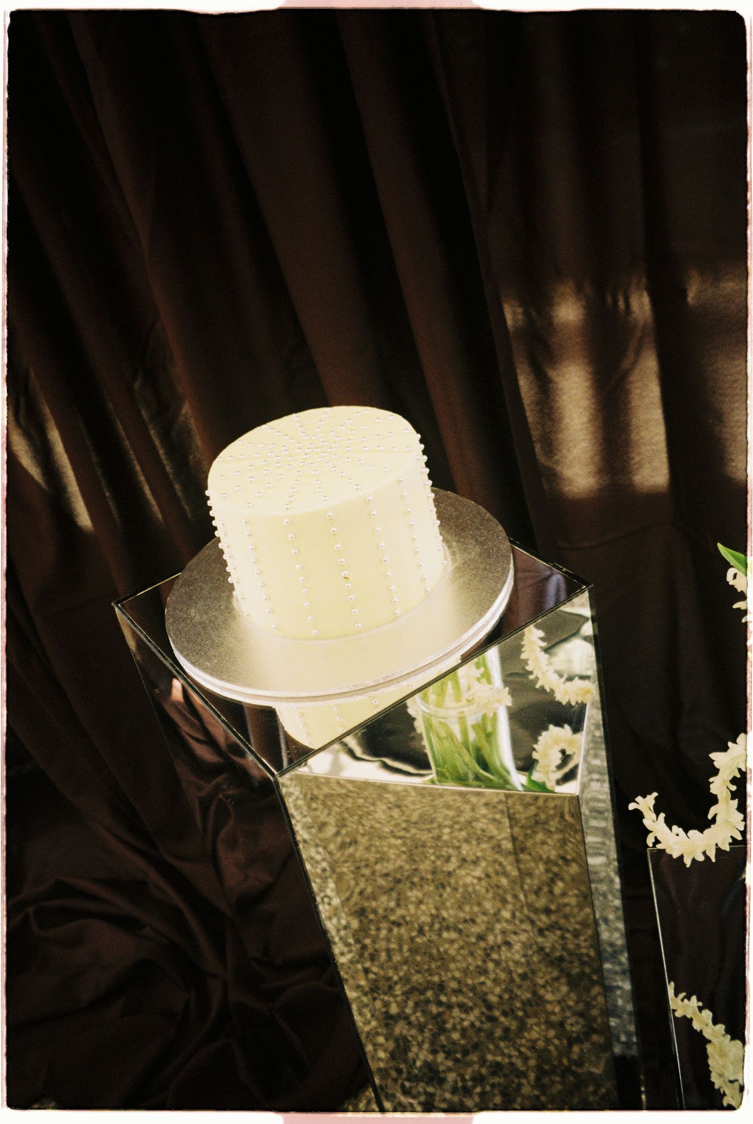 White single-tier wedding cake on mirrored pedestal with dark draped fabric backdrop.
