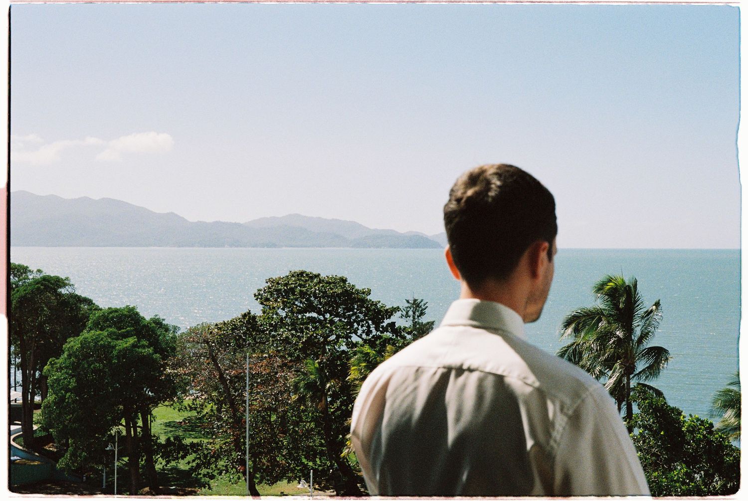 Man gazing at tropical ocean view with lush green trees, calm blue water, and misty mountains in the background.