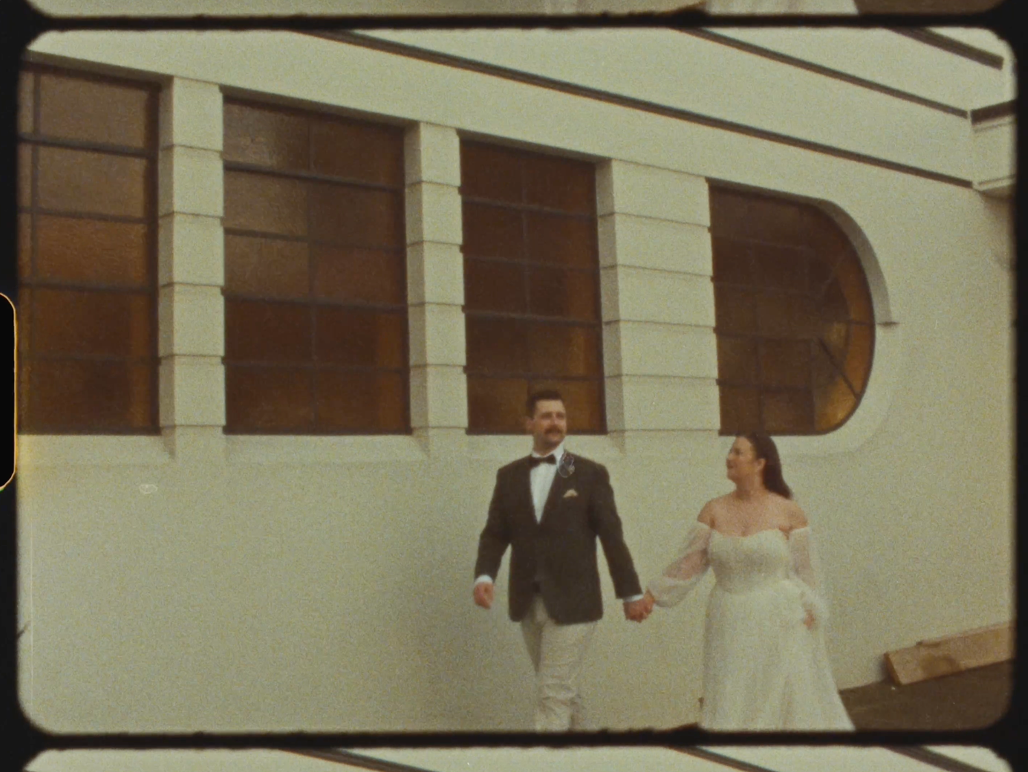 A bride and groom walk hand in hand outside a white Art Deco building.