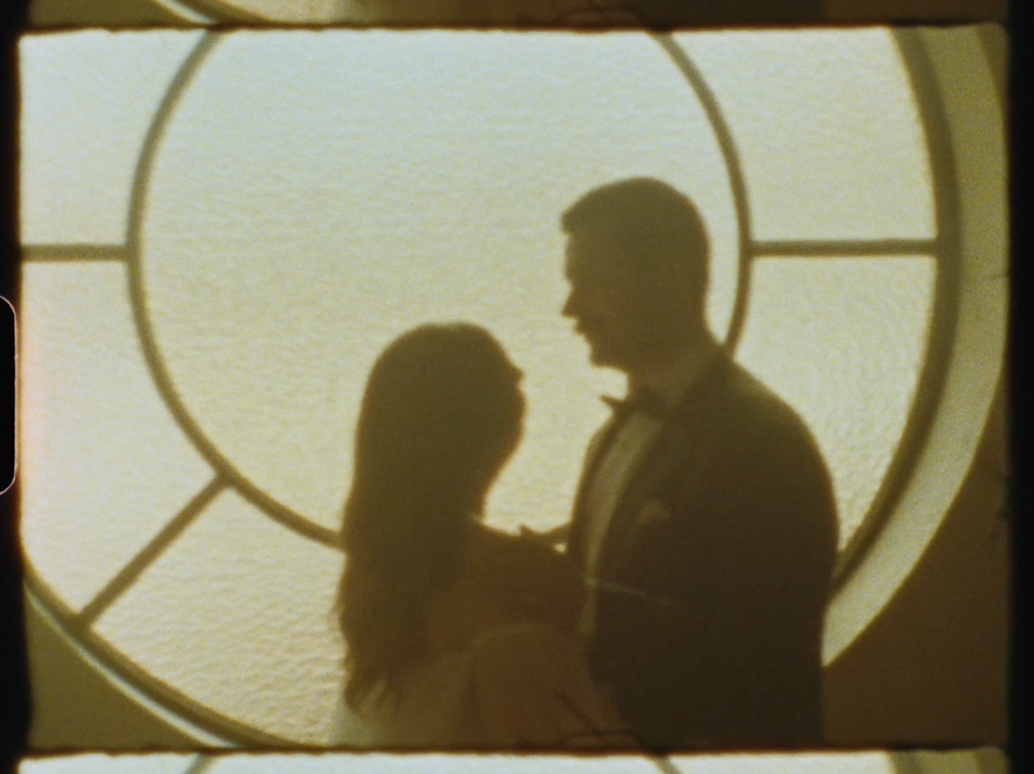 Silhouettes of a romantic couple facing each other framed by a circular window with soft golden light.