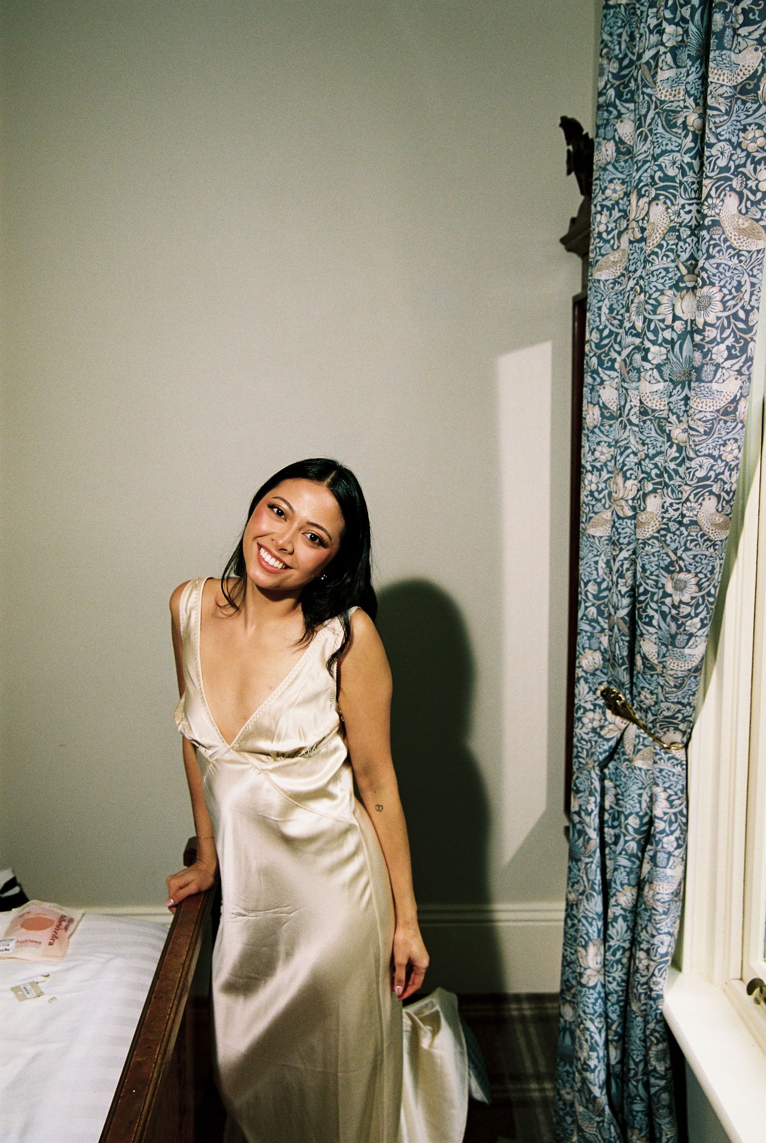 Bride smiling in elegant champagne satin slip dress standing by floral curtains indoors.