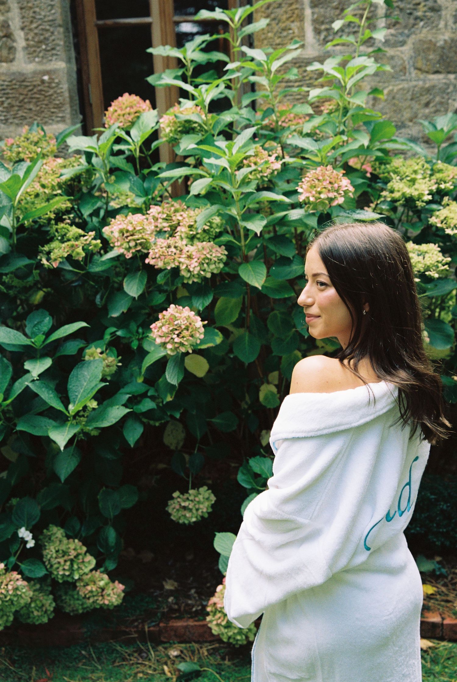Brunette woman in white off-shoulder robe stands beside blooming hydrangea bushes outdoors.