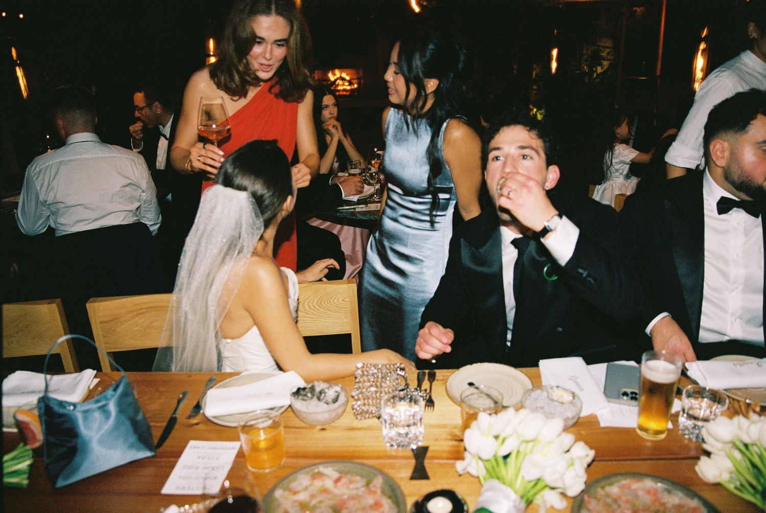 Bride in veil chats with tuxedo-clad groom and guests in formal attire at elegant wedding reception dinner.