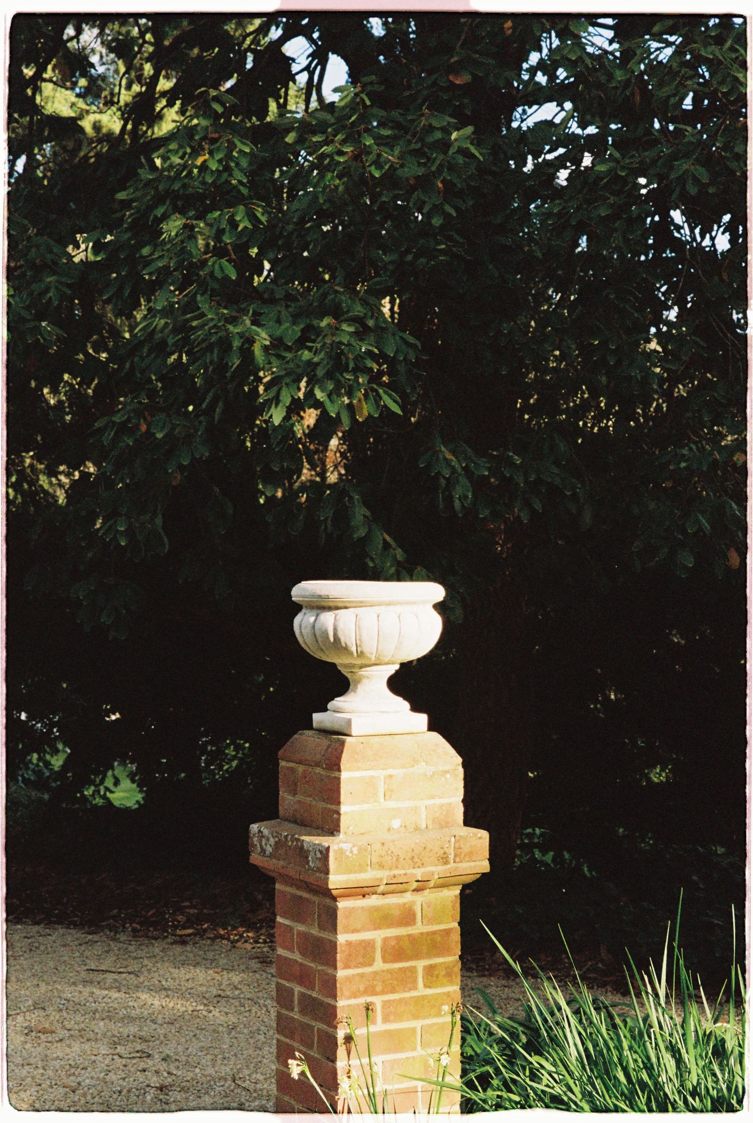 A decorative stone urn atop a classic brick pillar surrounded by lush green garden trees.