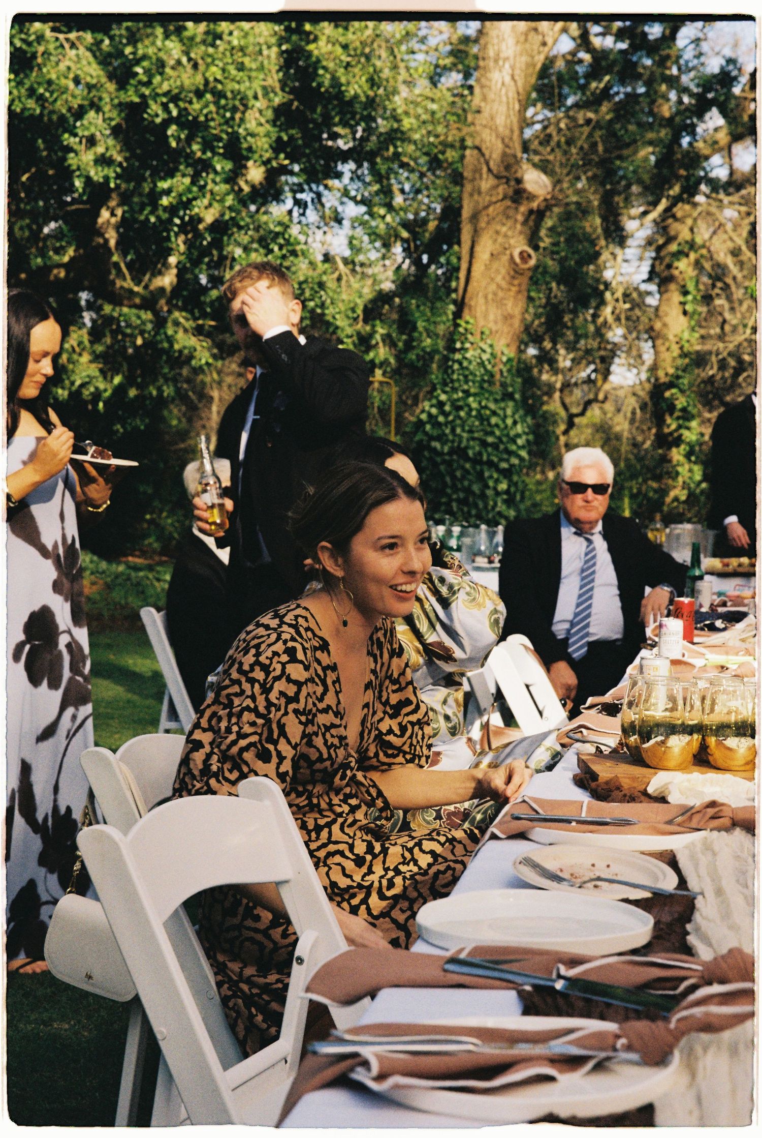 Elegantly dressed wedding guest smiles at outdoor garden reception table.