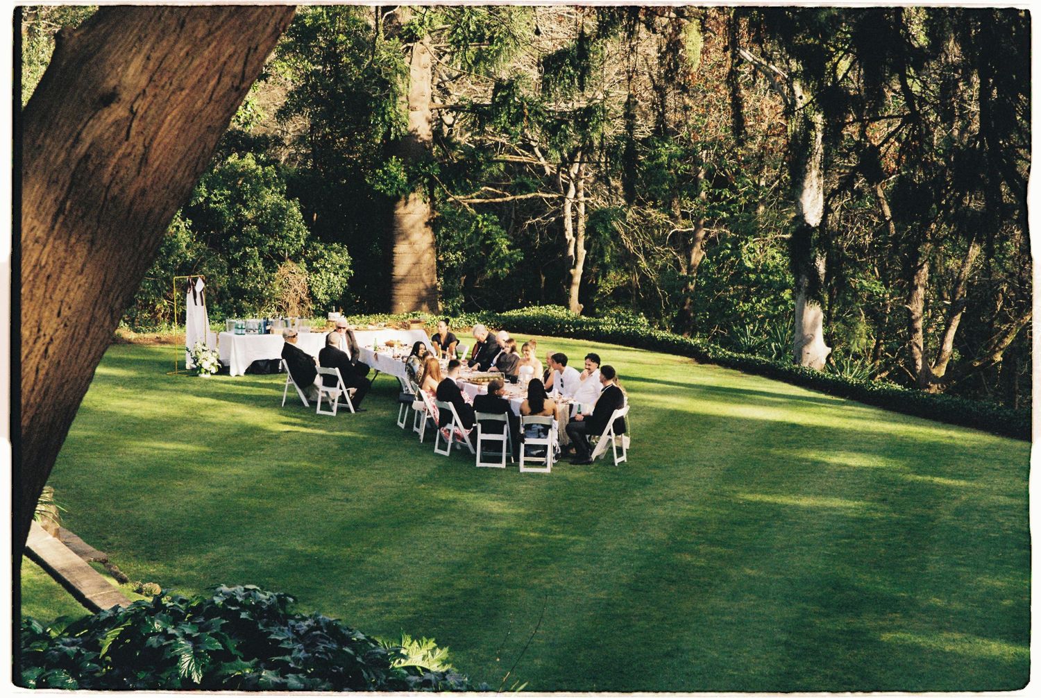 Outdoor wedding reception with guests seated at white tables on lush green lawn among trees.