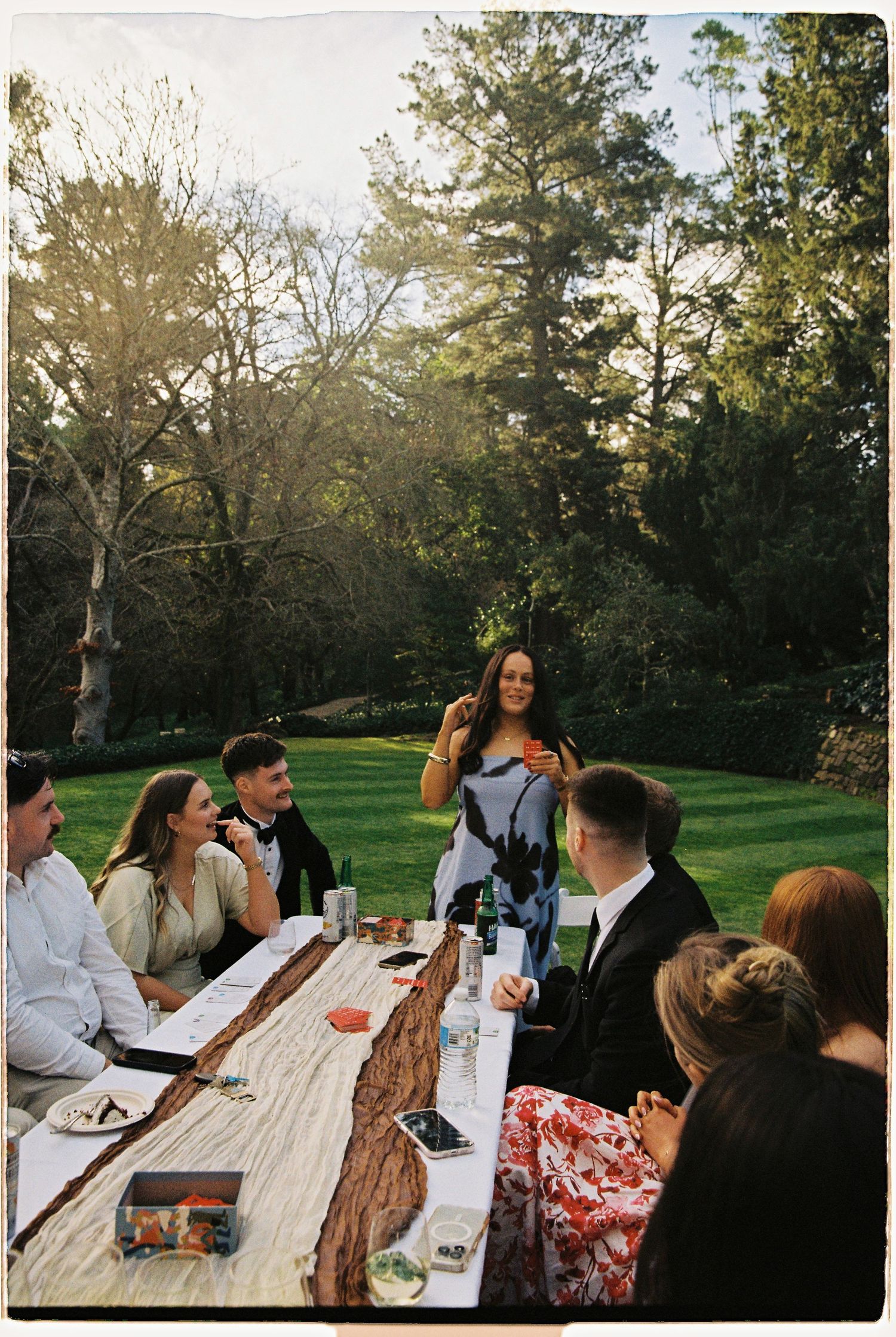 Wedding guest in floral dress giving a speech at an outdoor reception table in a sunlit garden.