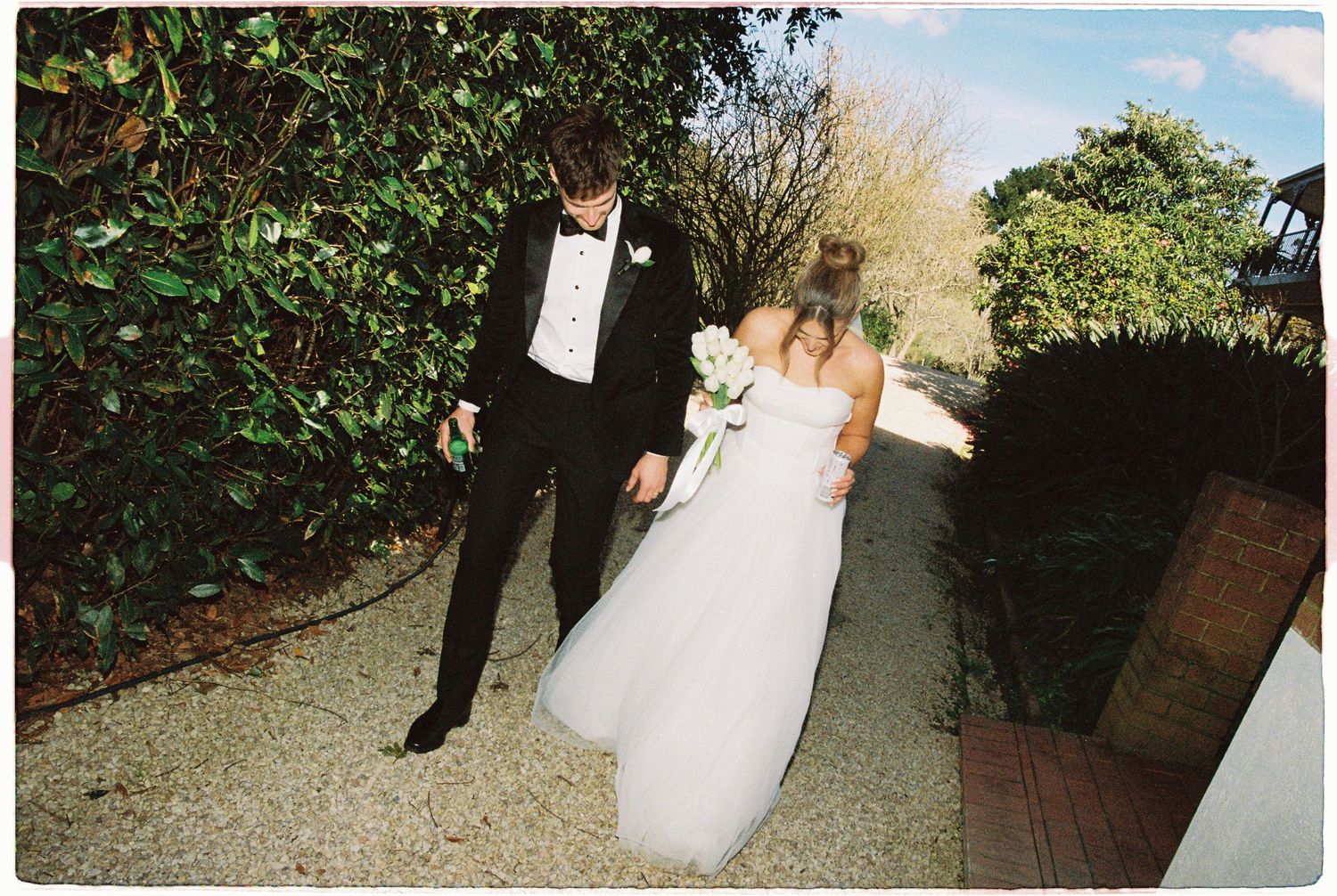 Bride in white tulle gown and groom in black tuxedo walk hand in hand along garden pathway.