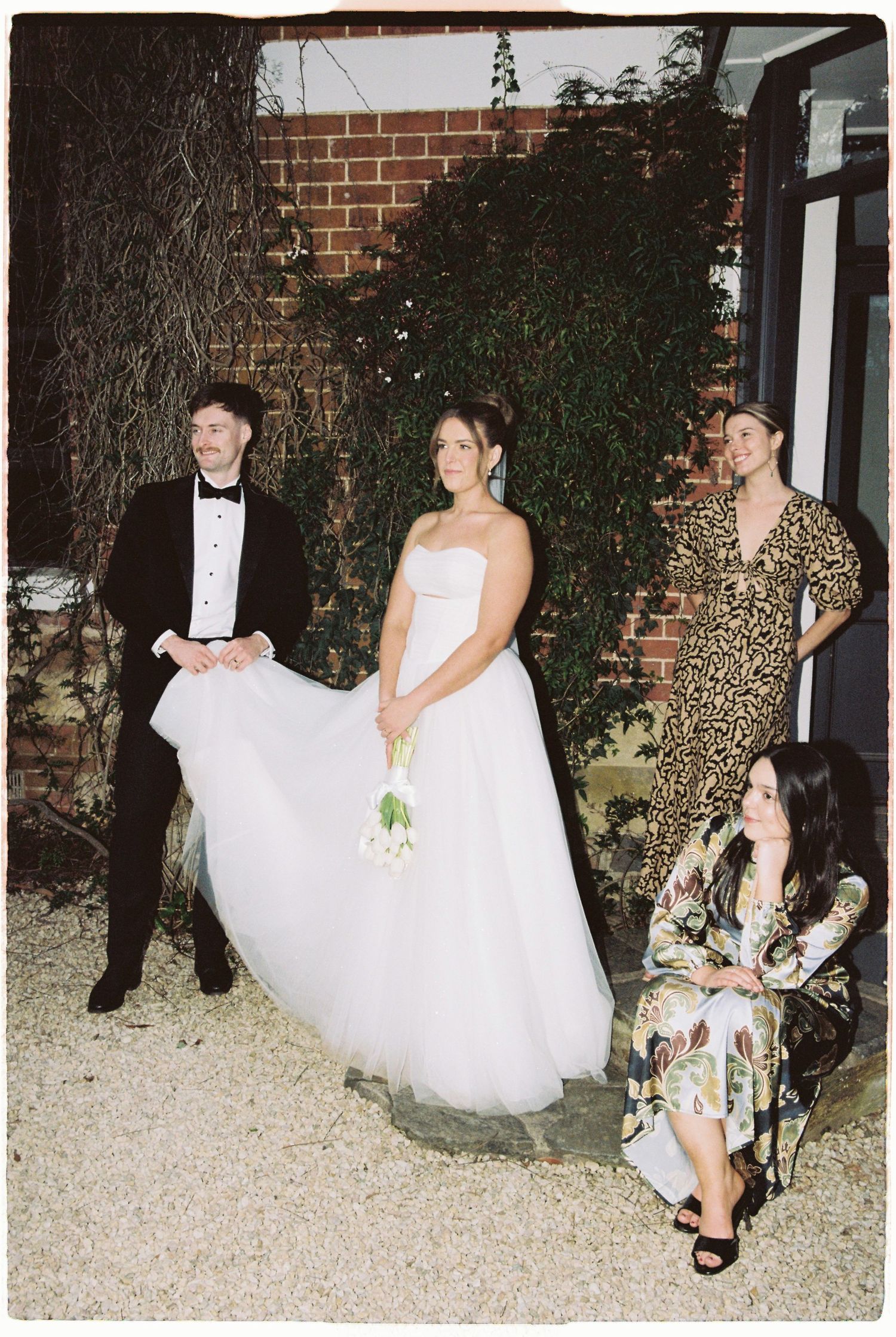 Bride in white gown poses with wedding guests in leopard print and floral attire outdoors.