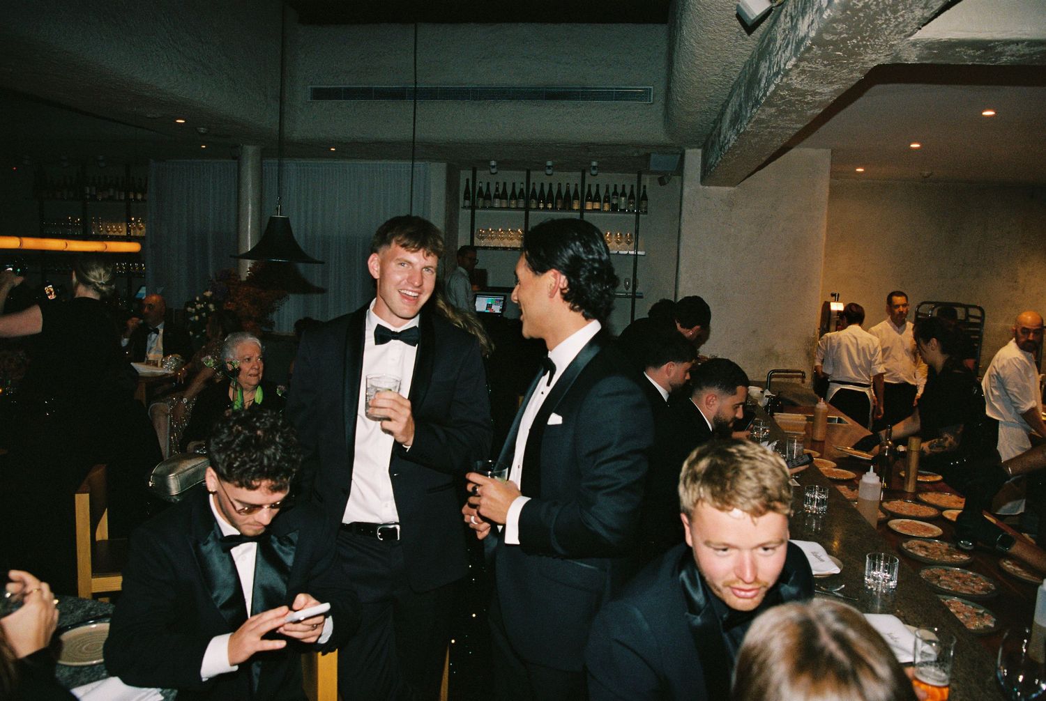 Elegantly dressed men in tuxedos socializing and laughing at a formal wedding reception.