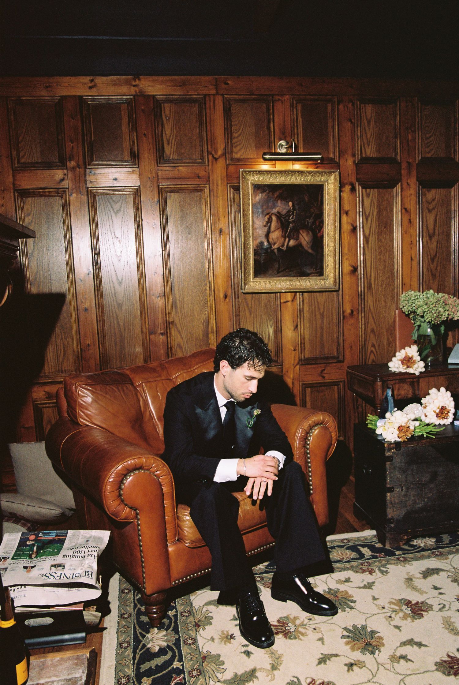 Groom in dark suit sitting pensively in leather armchair in wood-paneled room.