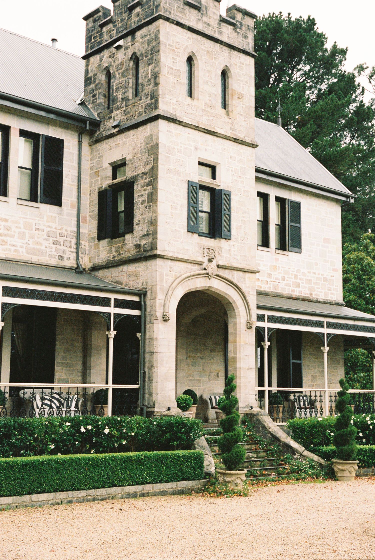 Historic stone castle-style manor with Gothic tower, black shutters, arched entry, and manicured topiary gardens.