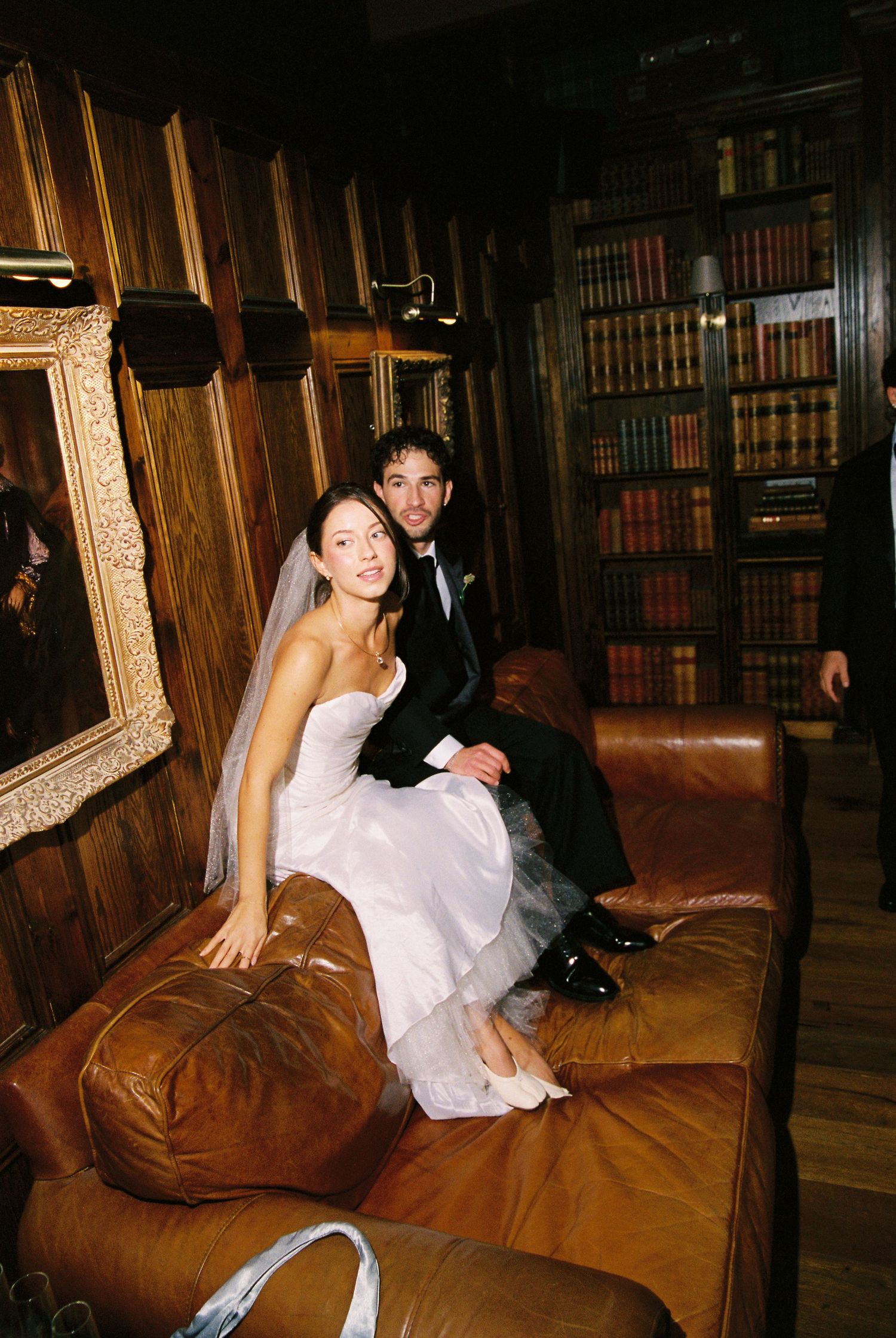 Bride and groom sitting together on leather sofa in wood-paneled library.