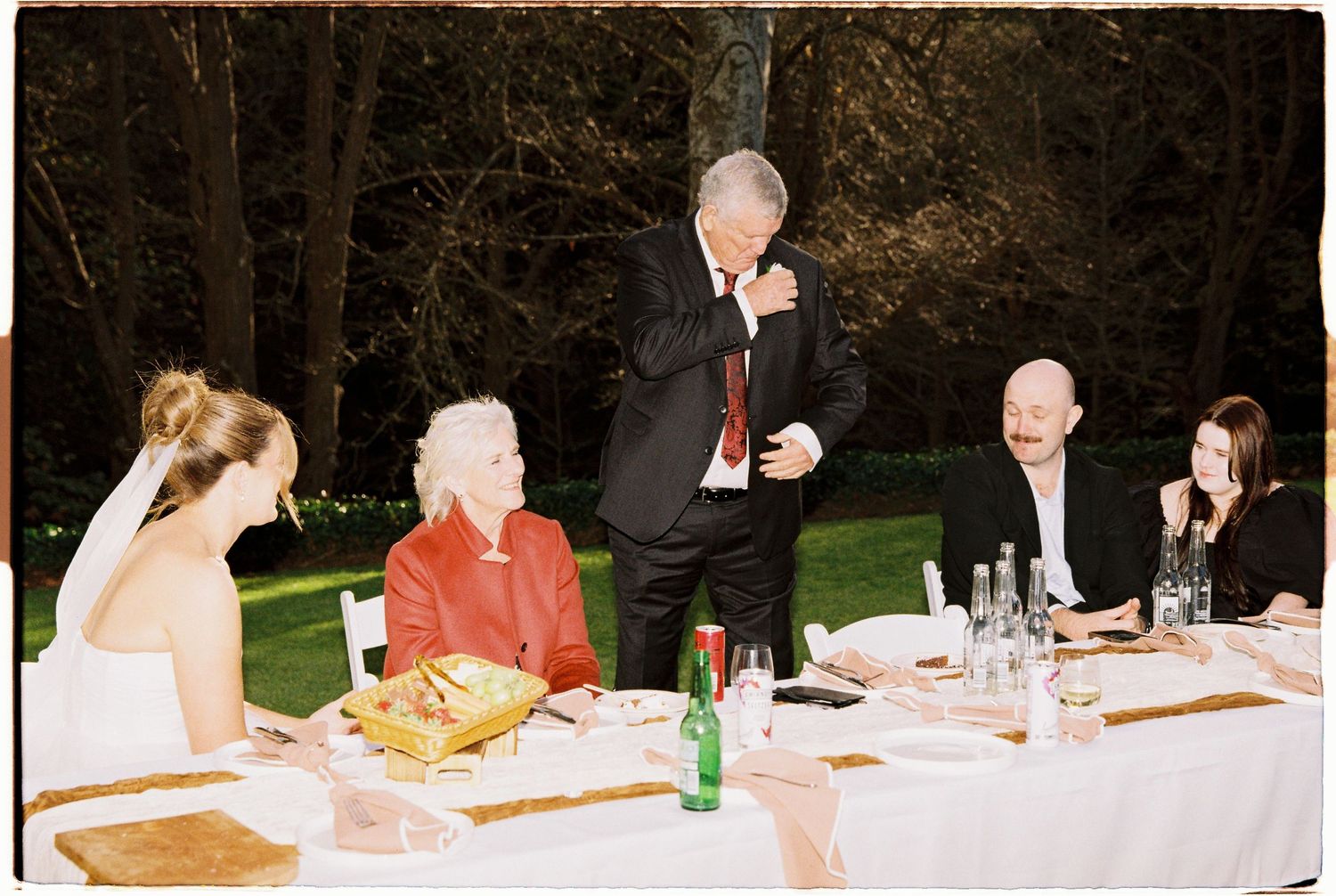 Elegant wedding reception dinner outdoors with guests seated at a decorated table as a man in a suit gives a heartfelt toast.
