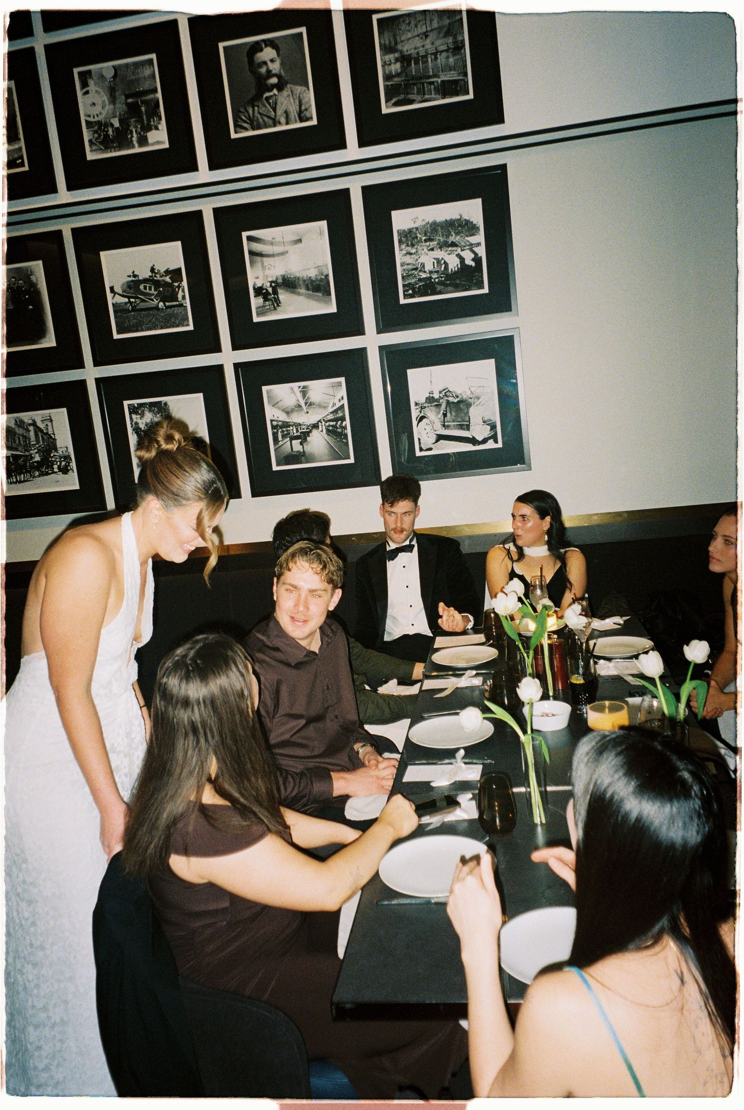 A bride in white mingles with guests seated at a elegantly set dinner table adorned with framed photos on the wall.