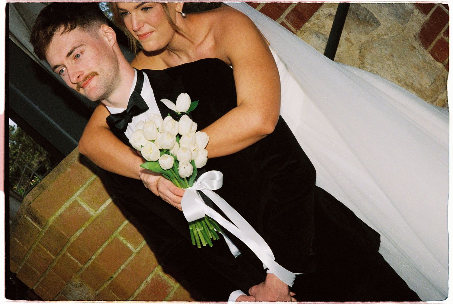 Bridesmaid in black gown holds white tulip bouquet alongside groomsman at wedding venue.